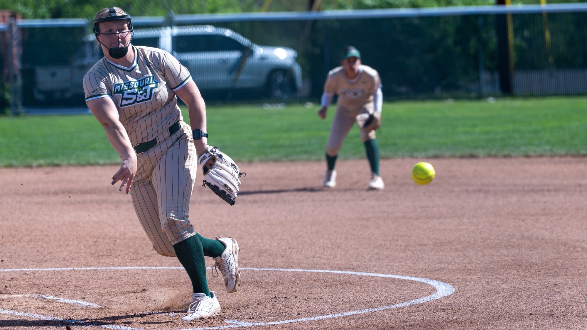 Mia Miller of the Missouri S&T Miners delivers a pitch during a softball game. She wears a pinstriped uniform and a protective pitcher's mask while releasing the ball from the circle.