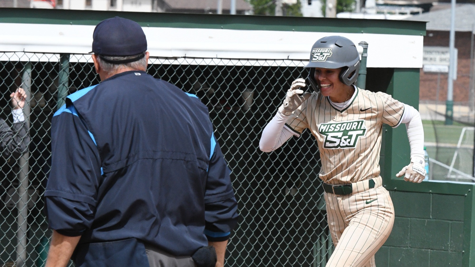 Missouri S&T Softball's Claire Lopez smiles as she runs the towards home plate after a home run. She is wearing a gold pinstripe softball uniform and dark gray batting helmet