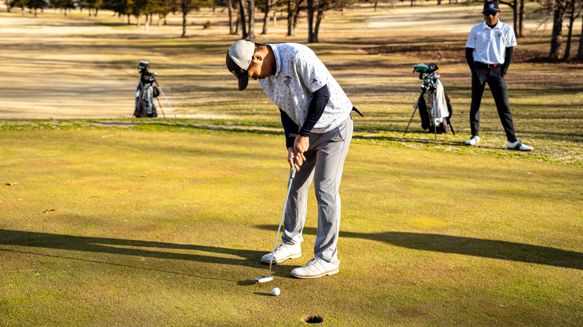 Isaac To of Missouri S&T golf is photographed lining up a short putt on a sunlit green, leaning forward with his putter as the ball sits a few feet from the hole, while a teammate and golf bags stand in the background among leafless trees.