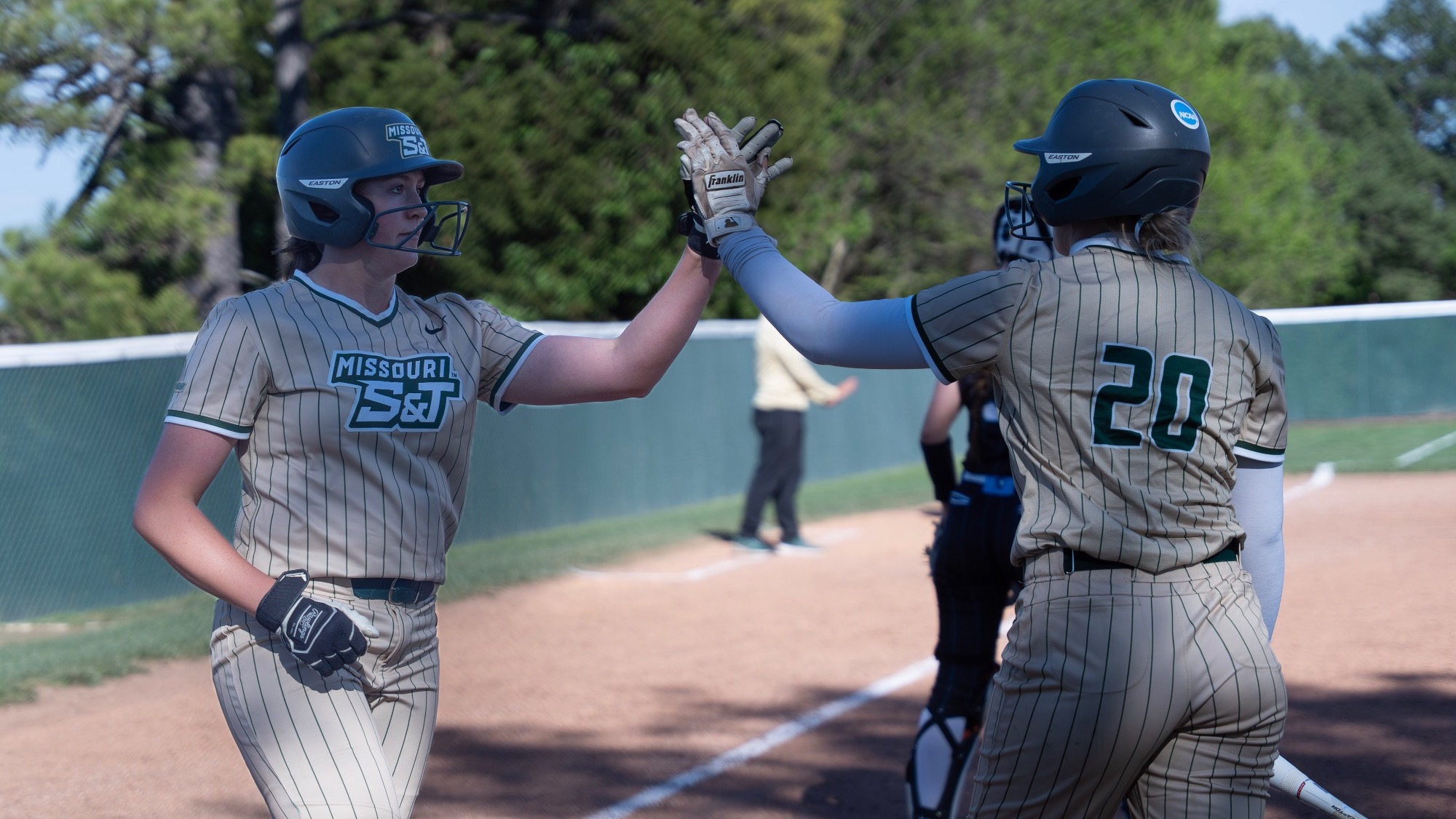 Tori Thompson (left) and Ty Neal (right) of Missouri S&T softball, both wearing tan pinstriped jerseys and helmets, high-five on the infield at the S&T softball field while celebrating a play.