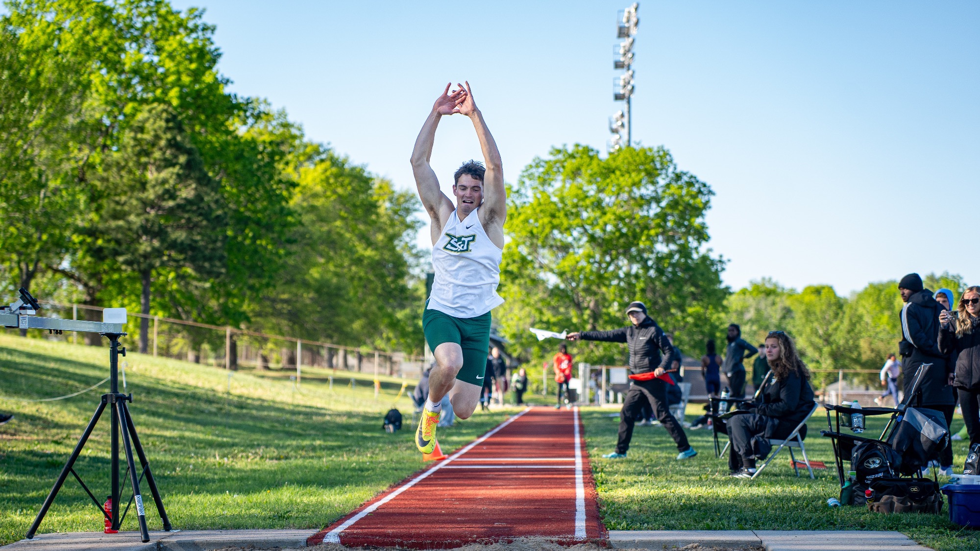 Missouri S&T’s Justin Chadwell competes in the triple jump at the Dewey Allgood Invitational, leaping into the sand pit with his arms extended overhead. He is wearing a white S&T track and field jersey with green shorts and bright spikes. In the background, the red runway stretches behind him with officials and spectators seated along the side, surrounded by green grass, trees, and clear skies.