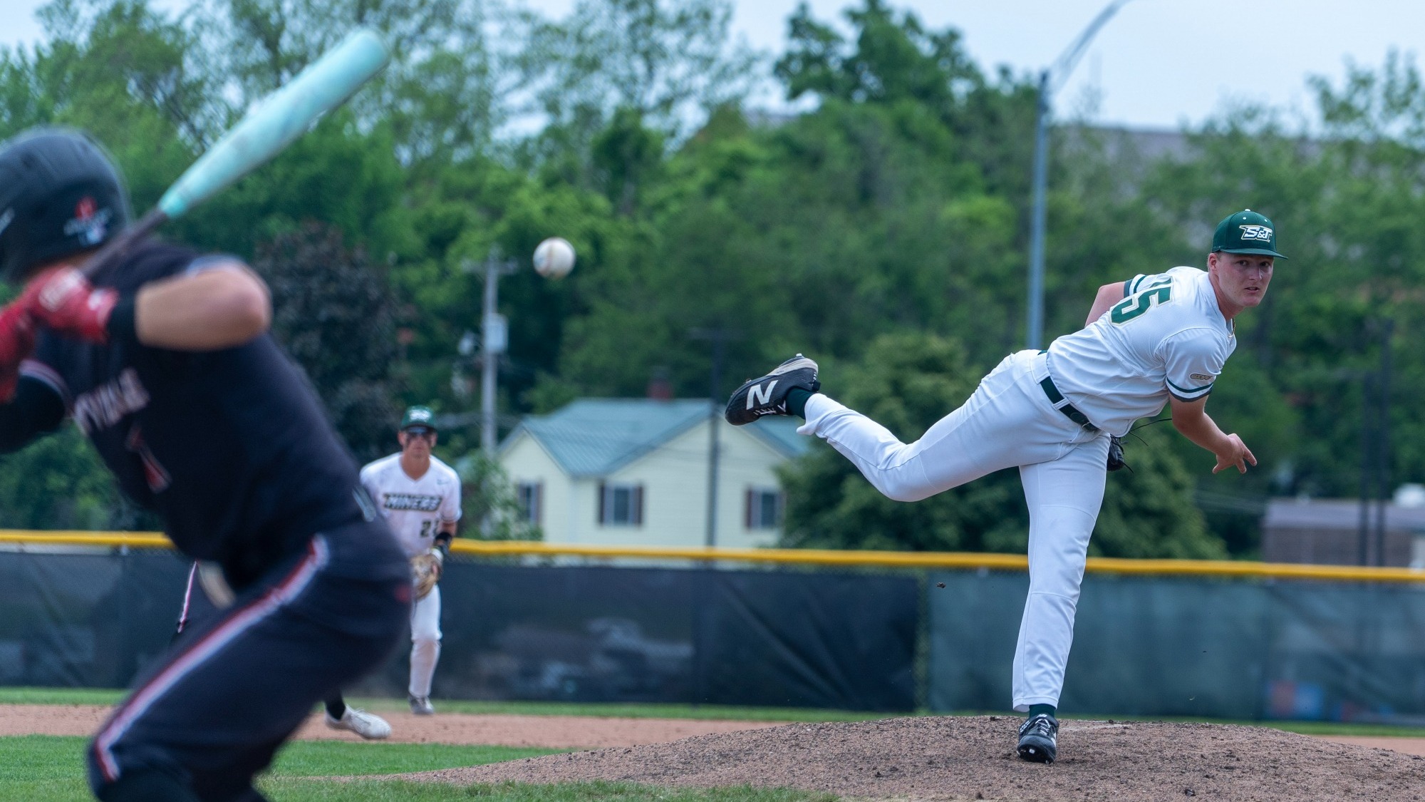Logan Slinkard of Missouri S&T baseball photographed during a pitch on the mound at the Ballpark at S&T. He has just released the ball and is on his follow through with the ball visible traveling through the air towards the batter. Slinkard is wearing a white jersey with green trim and a green baseball cap.
