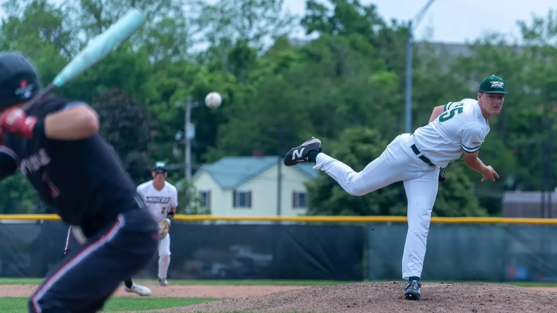 Logan Slinkard of Missouri S&T baseball photographed during a pitch on the mound at the Ballpark at S&T. He has just released the ball and is on his follow through with the ball visible traveling through the air towards the batter. Slinkard is wearing a white jersey with green trim and a green baseball cap.