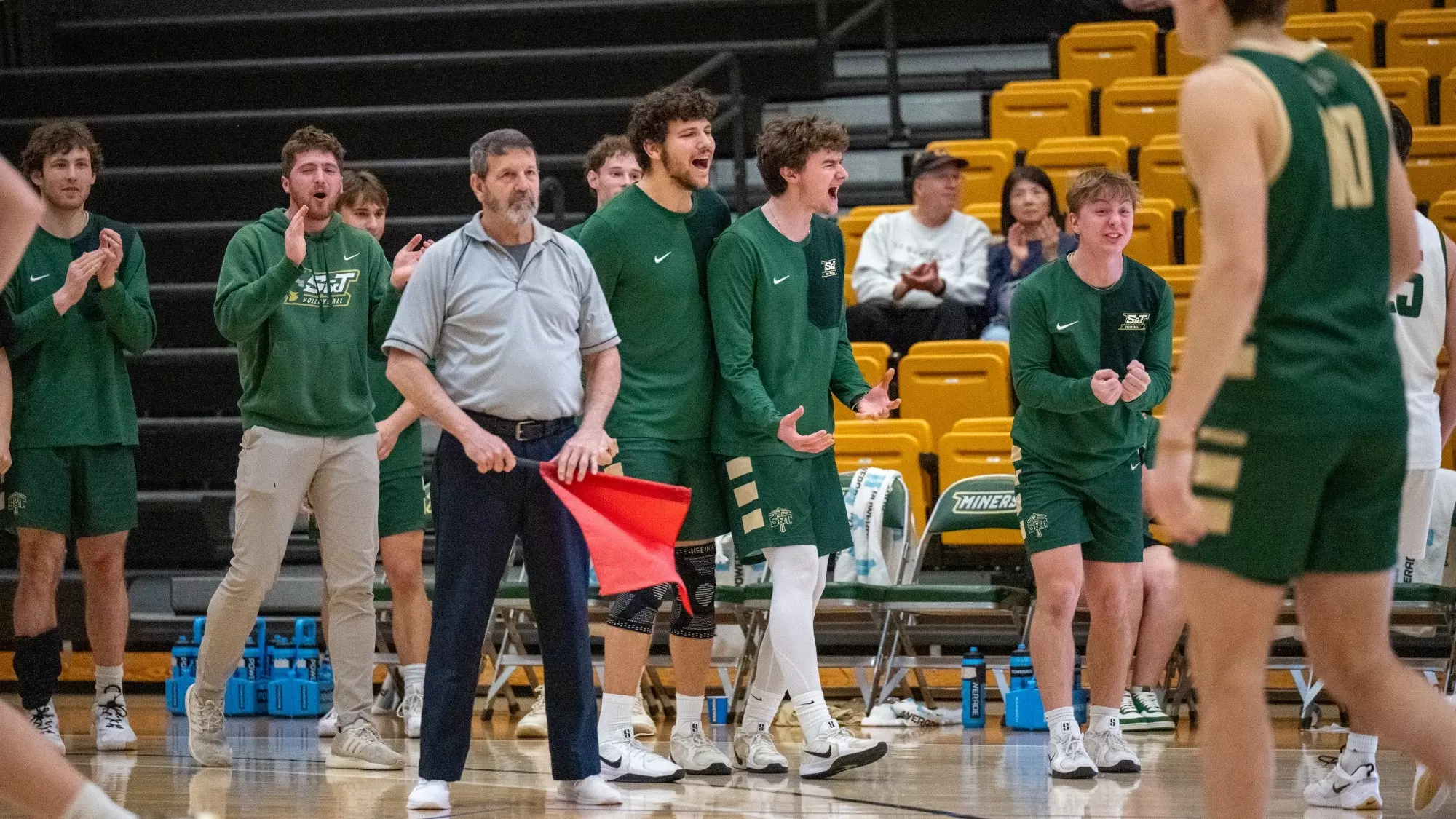 Missouri S&T men’s volleyball players in green warm-ups stand and react from the bench during a match, clapping and gesturing as play continues on the court, with arena seating visible in the background