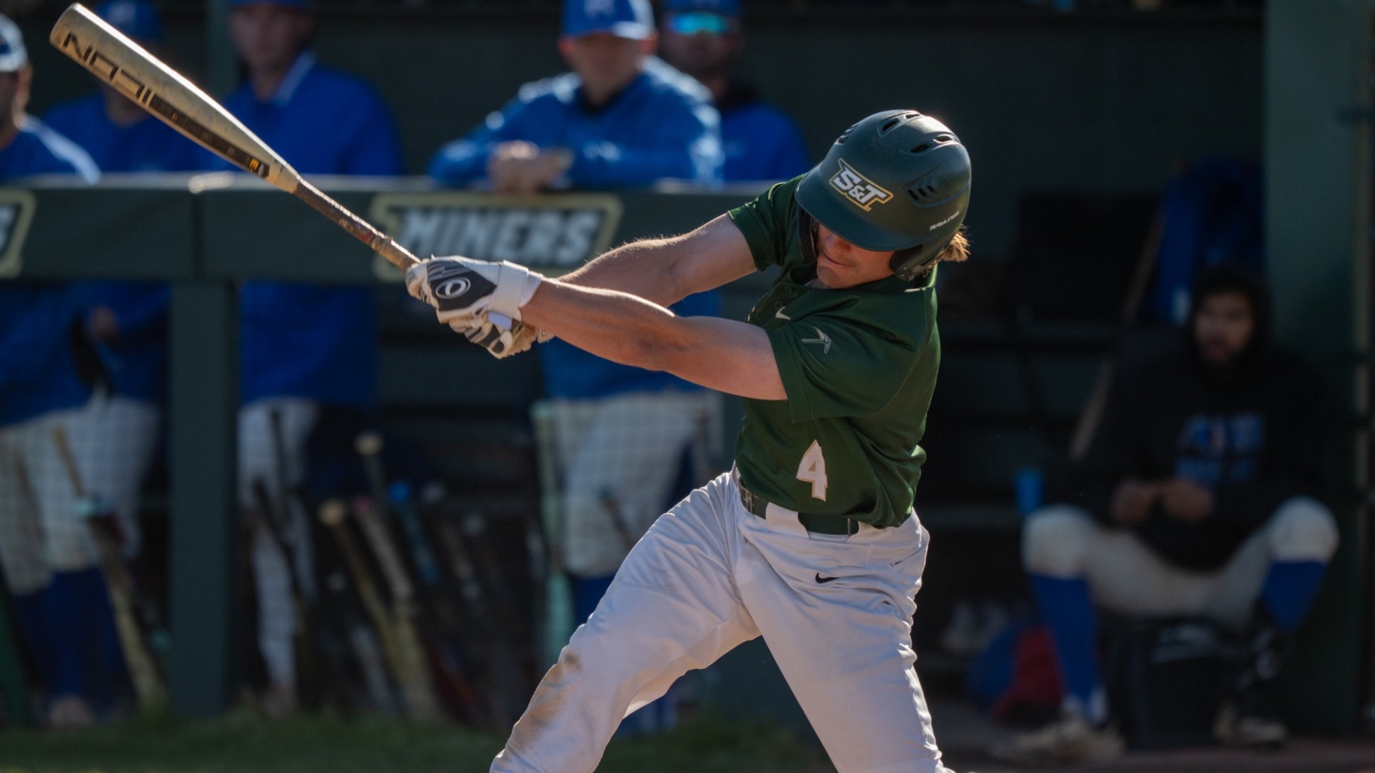 Aiden Cizek of the Missouri S&T Miners swings a bat during a game, wearing a green helmet and jersey with white pants in front of the dugout