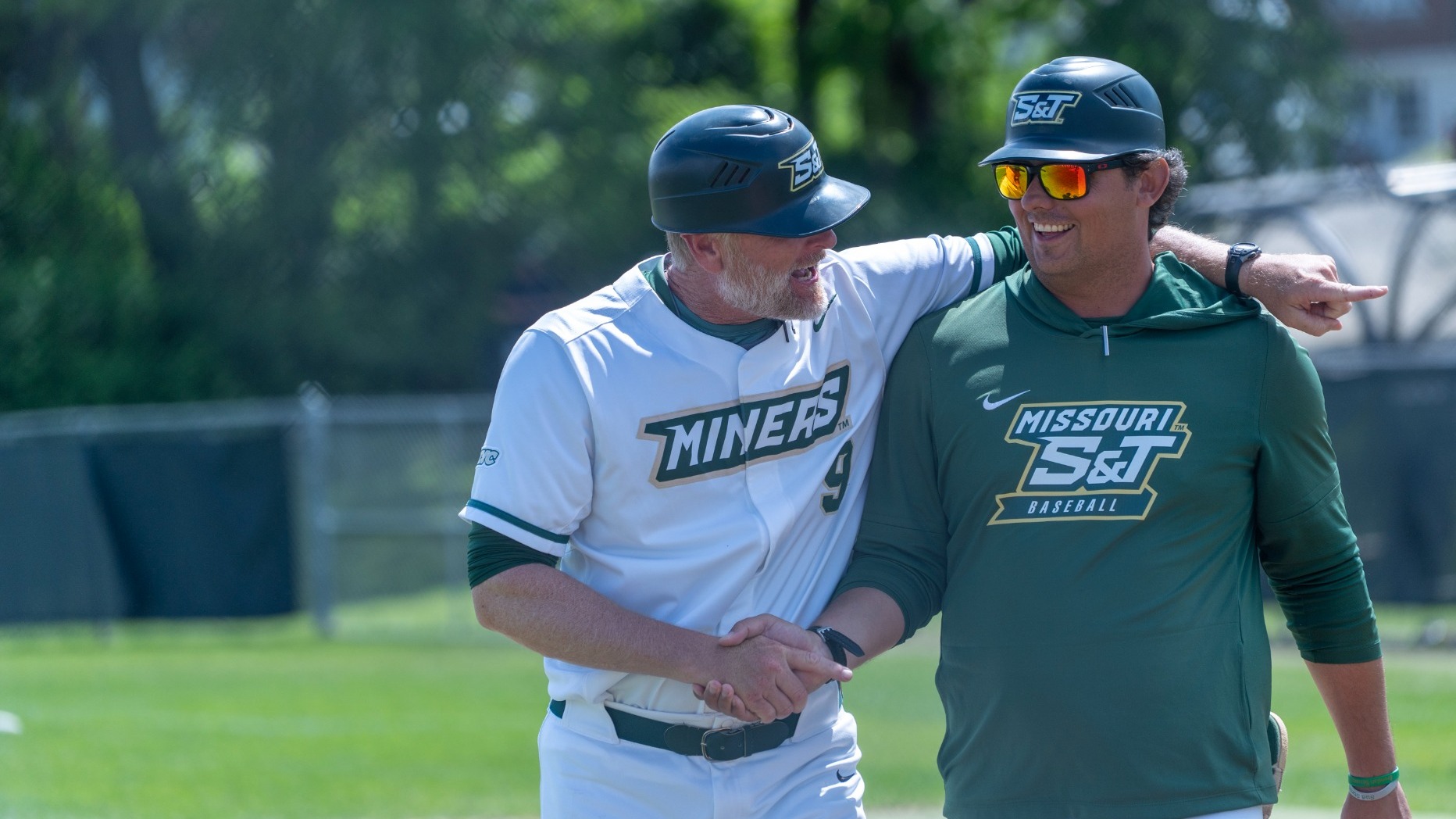 Baseball Head Coach Todd DeGraffenreid and Baseball Assistant Coaching Cody Gardner are photographed talking and smiling after a win over Maryville. They are shaking hands and walking side by side with Coach DeGraffenreid pointing and Coach Gardner smiling. DeGraffenreid wears a white S&T baseball uniform and Gardner wears a green shirt, sunglasses, and a hat