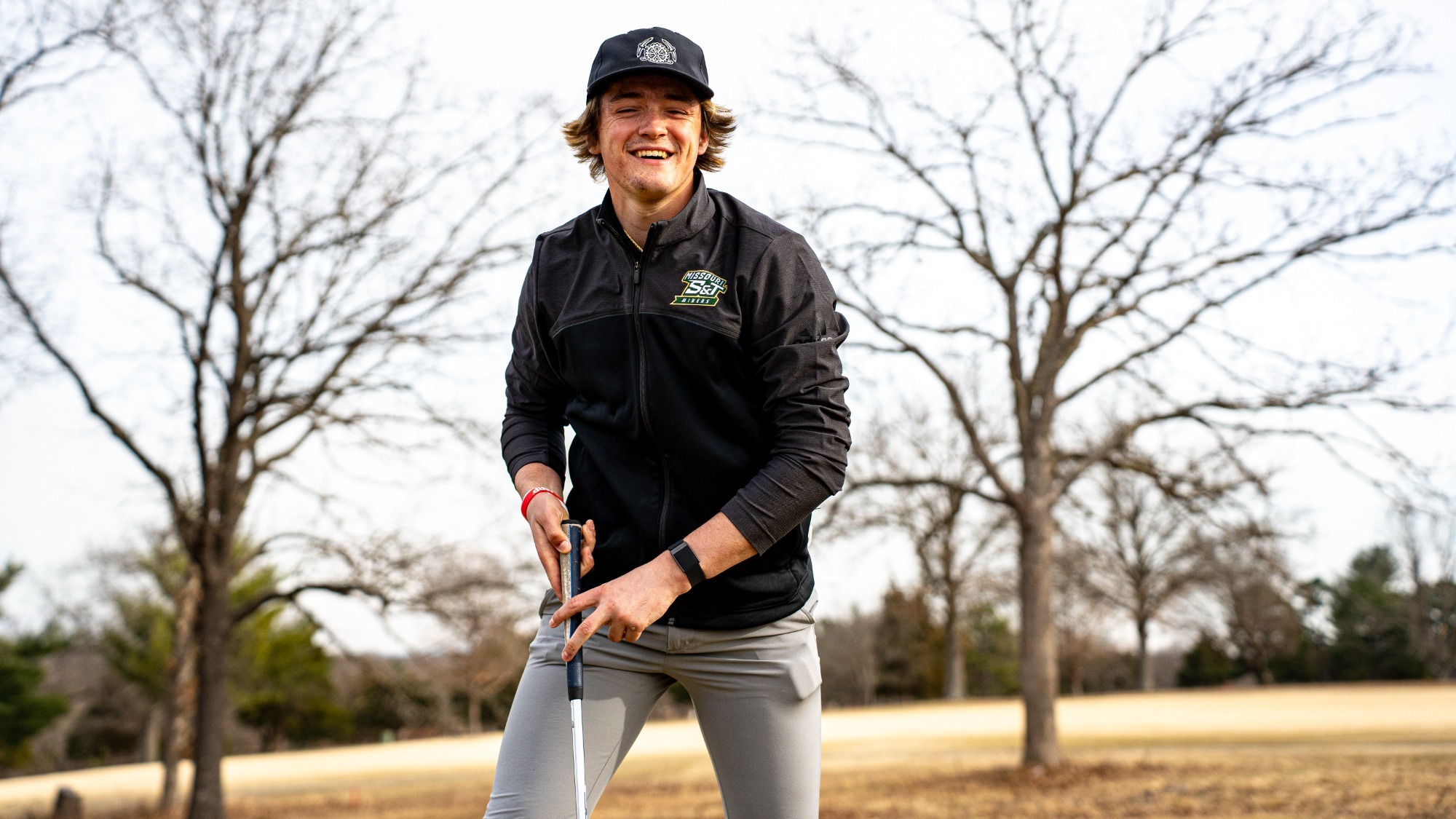 Chayton Jordan of the Missouri S&T Miners smiles while standing outdoors on a grassy field, holding a golf club with both hands as he prepares to take a shot, with leafless trees in the background