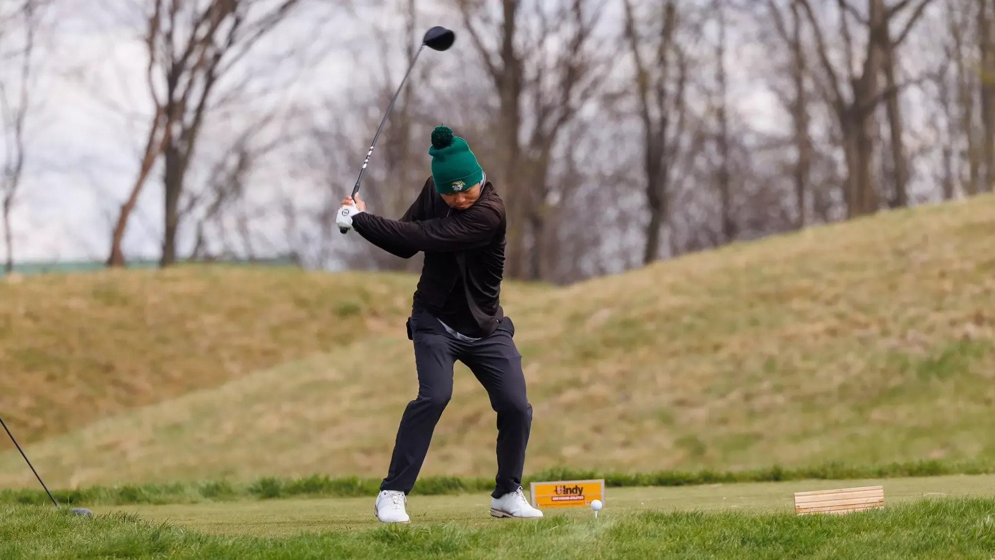 Isaac To swinging teeing off and swinging a driver during the Ken Partridge Invitational. He is wearing a green Miner knit cap with a black shirt and dark gray shorts. He is in the middle of his back swing and looking down at the golf ball.
