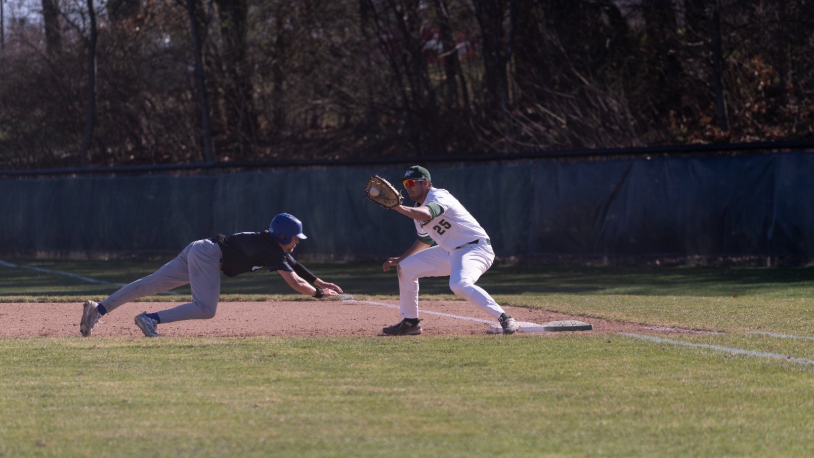 Nick Hooper, a Missouri S&T first baseman in a white uniform, stretches from first base with his glove extended to receive a throw as a Grand Valley State baserunner in a dark jersey dives headfirst back to the bag. The play takes place on the infield dirt near the foul line, with the outfield fence and trees visible in the background, capturing a close pickoff attempt at first base during daylight action