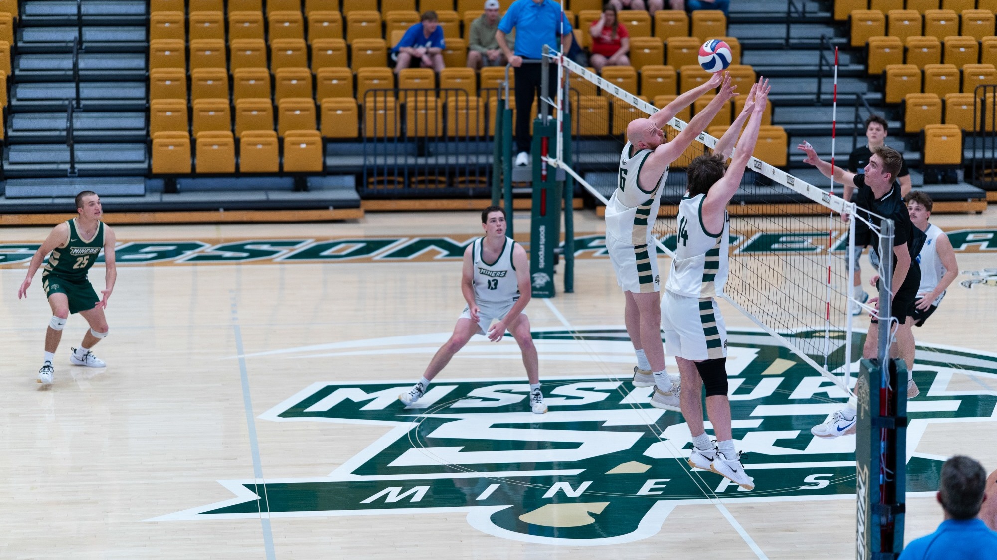 Missouri S&T volleyball players #6 and #14 jump together at the net to block an opposing hitter in a black jersey during a rally at Gibson Arena, while teammates #13 and #23 prepare defensively behind them on the Miners logo at center court