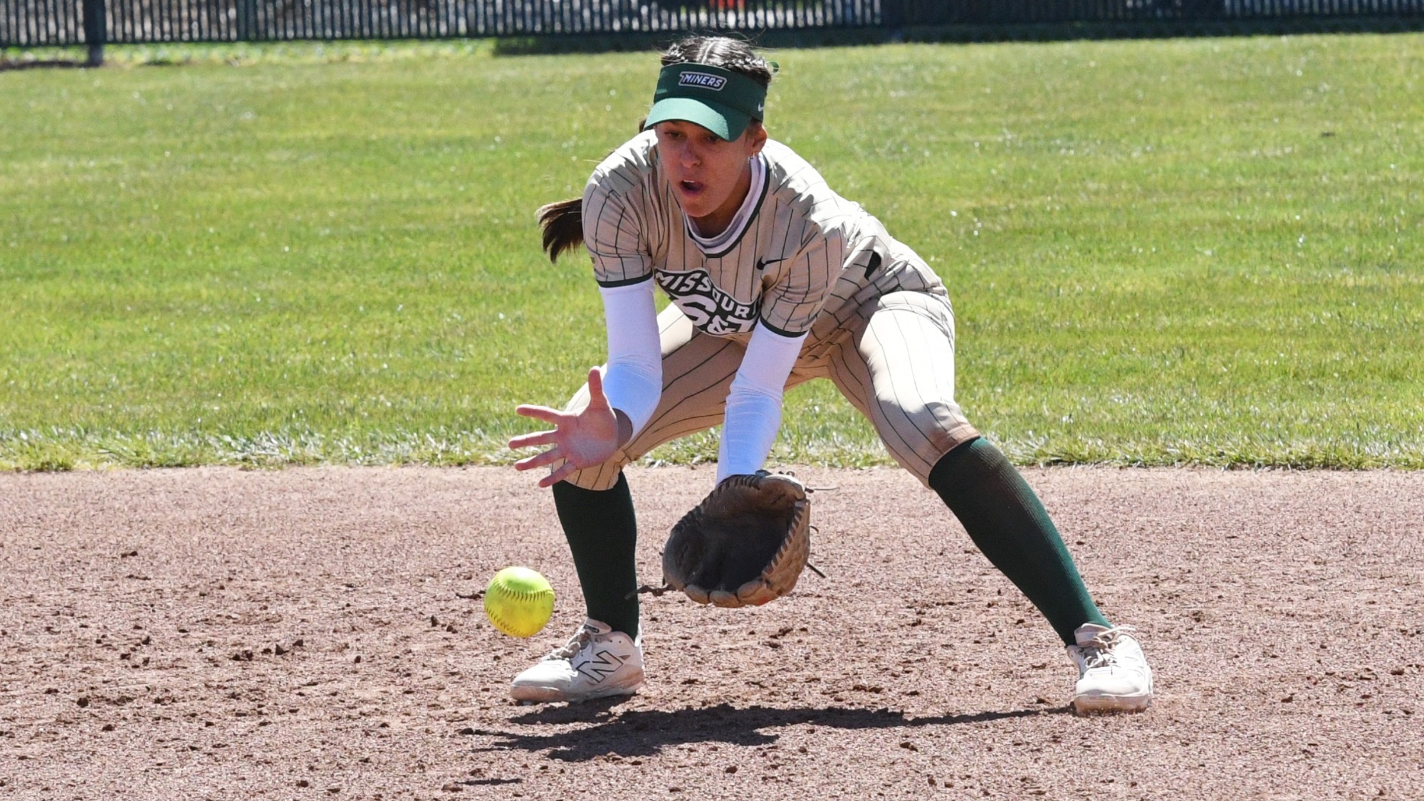 Missouri S&T second baseman Claire Lopez fields a ground ball on the infield dirt, reaching forward with her glove while wearing a gold pinstripe Miners jersey at the Missouri S&T softball field.
