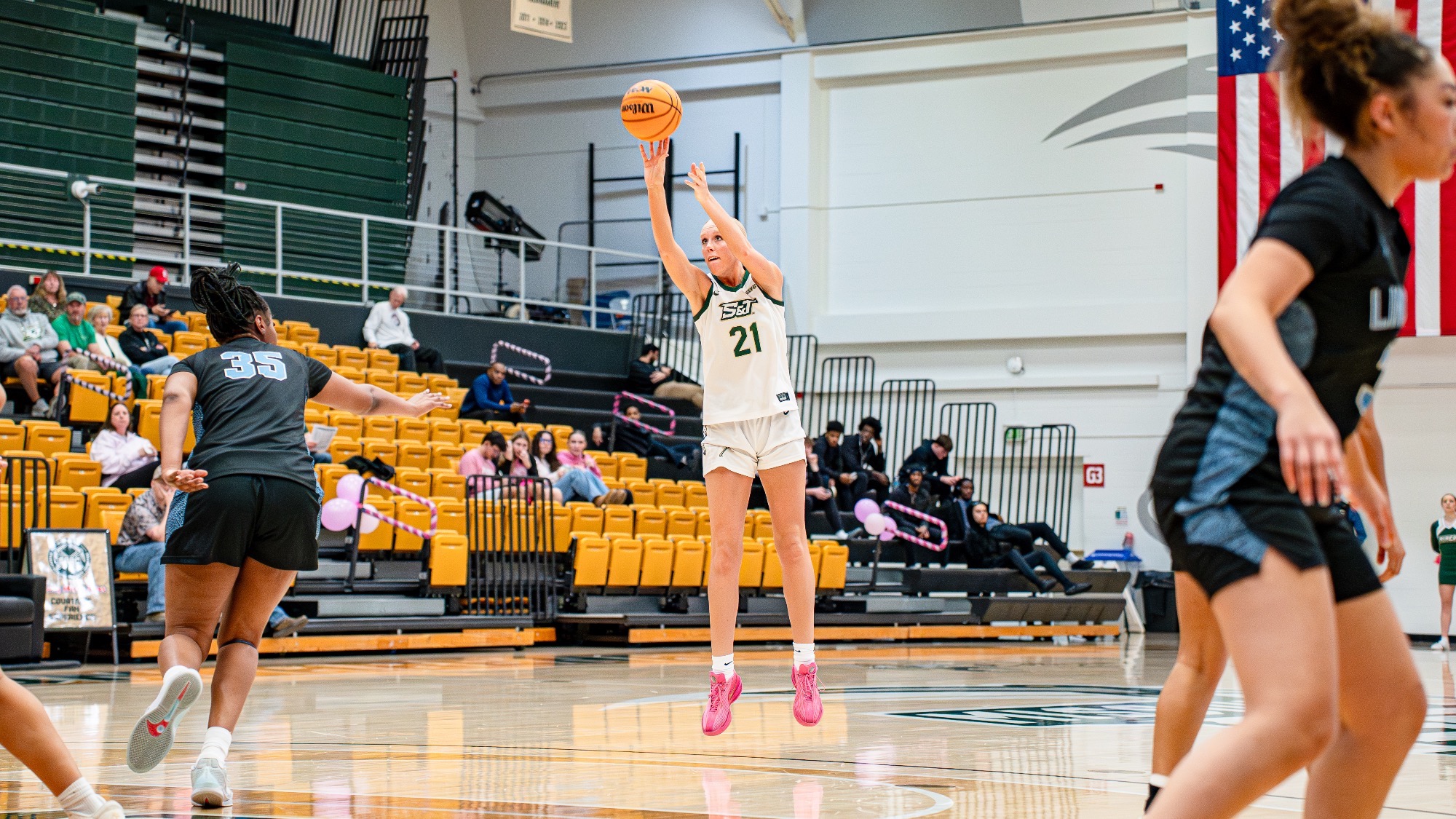 Missouri S&T senior forward Morgan Luebbering, wearing a white Miners jersey, elevates to shoot a three-pointer from the top of the key in Gibson Arena as defenders close in.