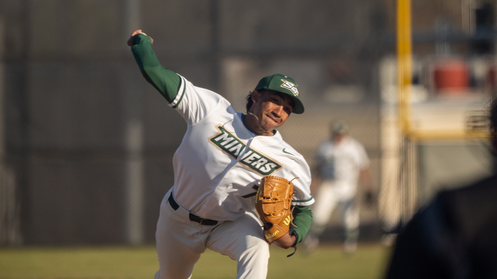 Dylan Wipf, a Missouri S&T Miners baseball pitcher in a white uniform with green sleeves and cap, delivers a pitch from the mound during a game, his throwing arm extended forward as an umpire stands blurred in the foreground and a teammate is visible in the background.