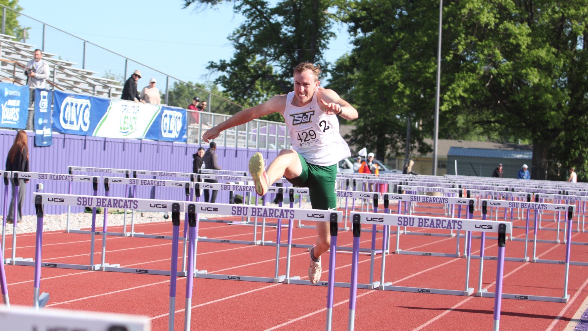 Kael Knittel of Missouri S&T, wearing a white S&T jersey and green shorts, clears a hurdle during the 110m hurdles (decathlon) at the GLVC Outdoor Championships at Southwest Baptist University, with hurdles and spectators visible in the background.