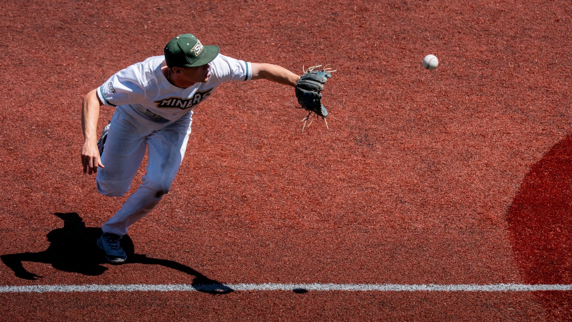 Will Muench of Missouri S&T Baseball team moves to his left with his baseball gloved hand extended towards a baseball to field it. He is wearing a white uniform and green baseball cap.