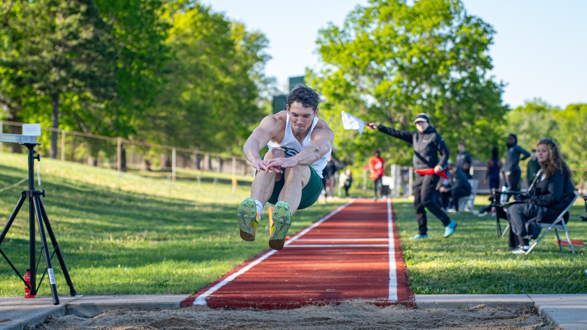 Justin Chadwell of Missouri S&T competes in the triple jump during a track and field meet, soaring through the air above the sand pit while wearing a white S&T singlet and green shorts. An official waves a white flag beside the runway as spectators watch in the background.
