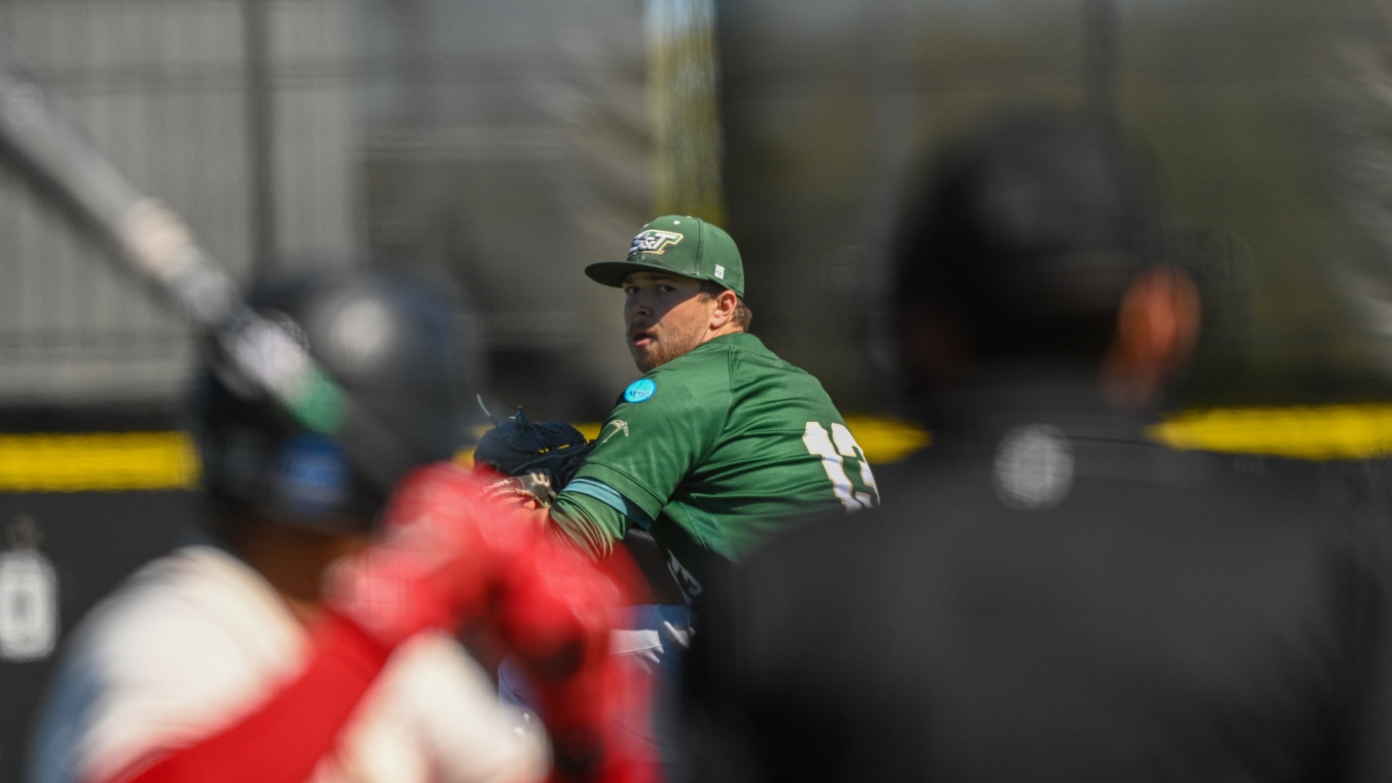 Dylan Bates of Missouri S&T Baseball photographed during a game at Grand Valley, he is looking at the batter and umpire who are blurred in the foreground as he gets ready to deliver a pitch