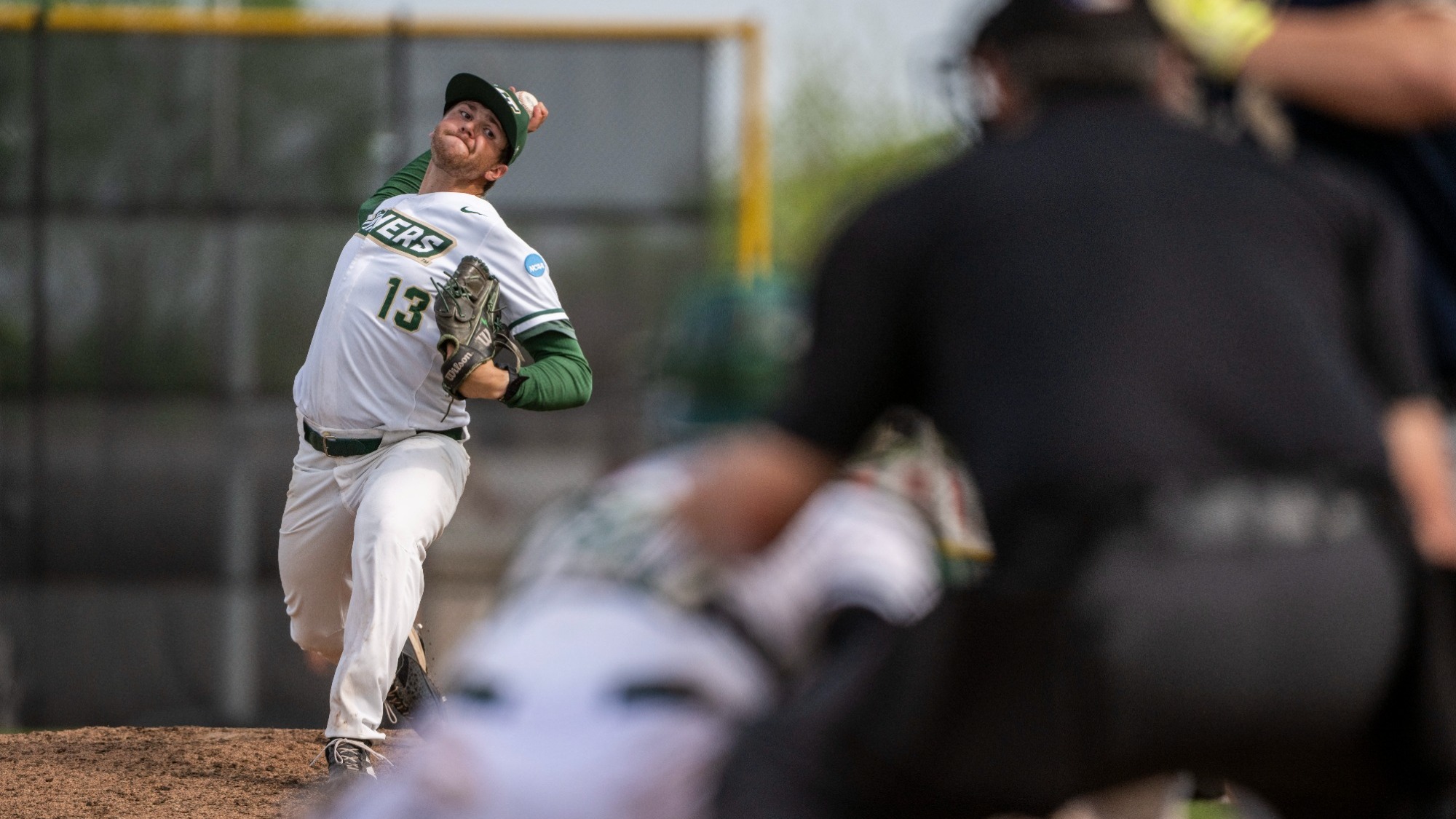 Dylan Bates of Missouri S&T Baseball photographed wearing a white uniform with green trim and lettering. He is in the process of delivering a pitch during a game at the ballpark at S&T with his right arm behind his head as he gets ready to throw. The batter, umpire, and catcher are blurred in the foreground