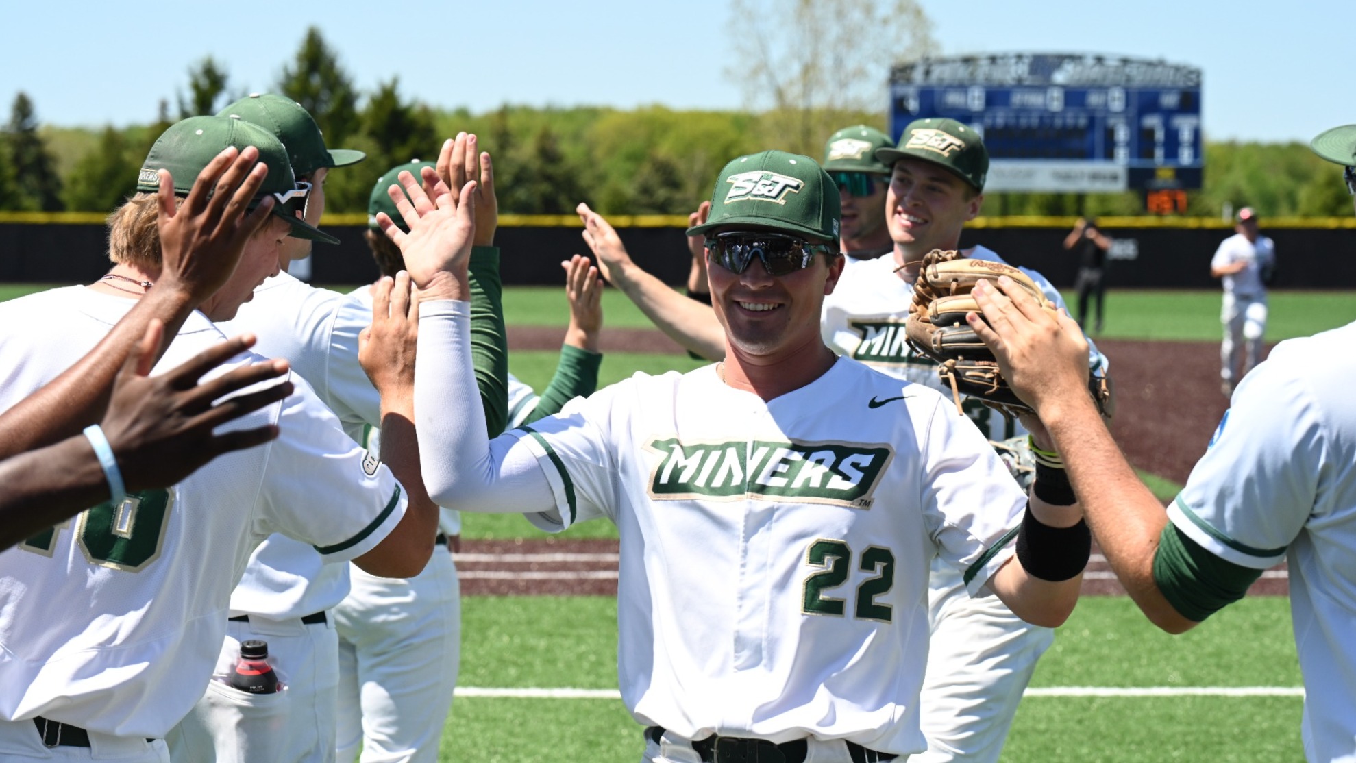 Nick Krodinger of Missouri S&T photographed smiling and high fiving teammates after successfully getting an out in the field. He is wearing an all white uniform with Green and Gold lettering and trim with a green baseball cap and sunglasses