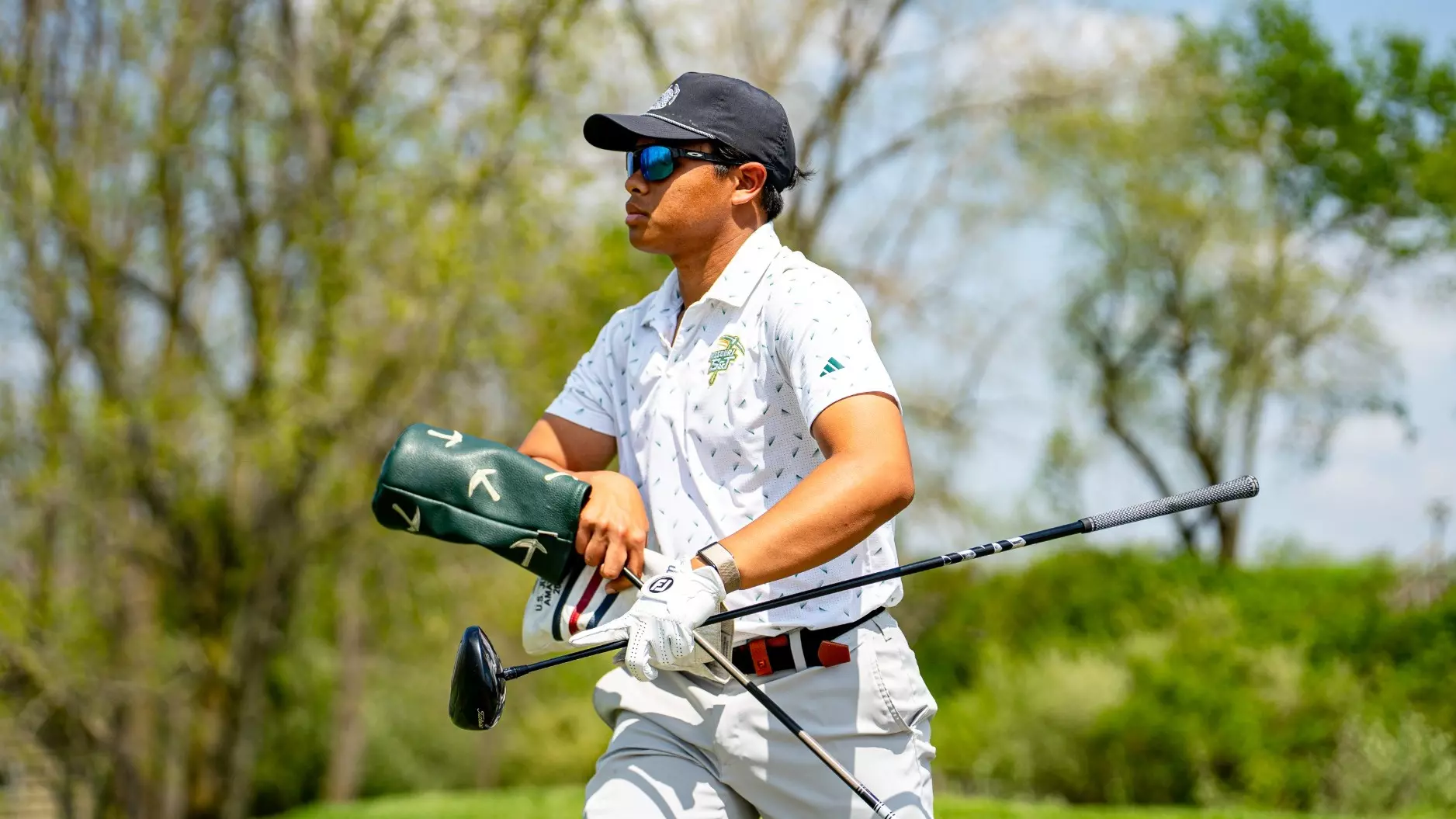 Rizq Adam Rohizam walks across the golf course carrying multiple clubs during competition, photographed wearing a Missouri S&T polo, sunglasses, and a black cap with trees and blue sky in the background.