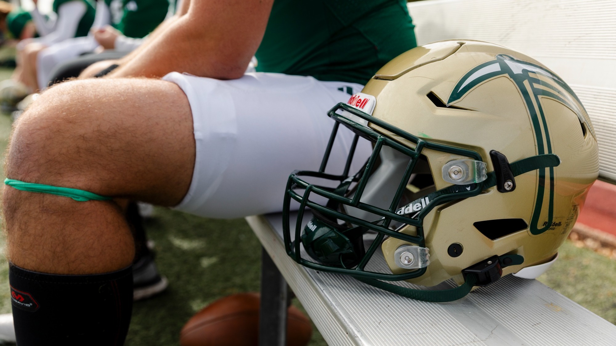 Football helmet resting on a sideline bench beside a seated Missouri S&T player in green and white practice gear, with teammates blurred in the background during an outdoor practice.
