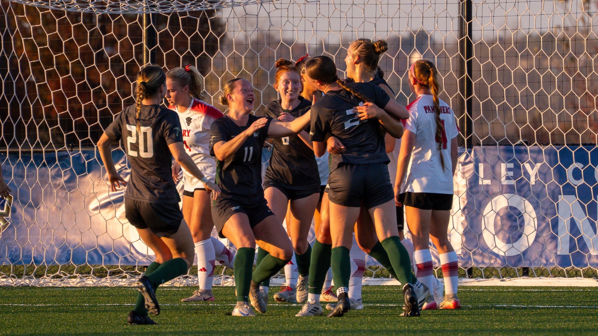 Missouri S&T women’s soccer players celebrate a goal during the GLVC Tournament in St. Louis, gathering in front of the net while wearing grey jerseys, grey shorts, and green socks as opposing players in white and red uniforms stand nearby during sunset conditions.