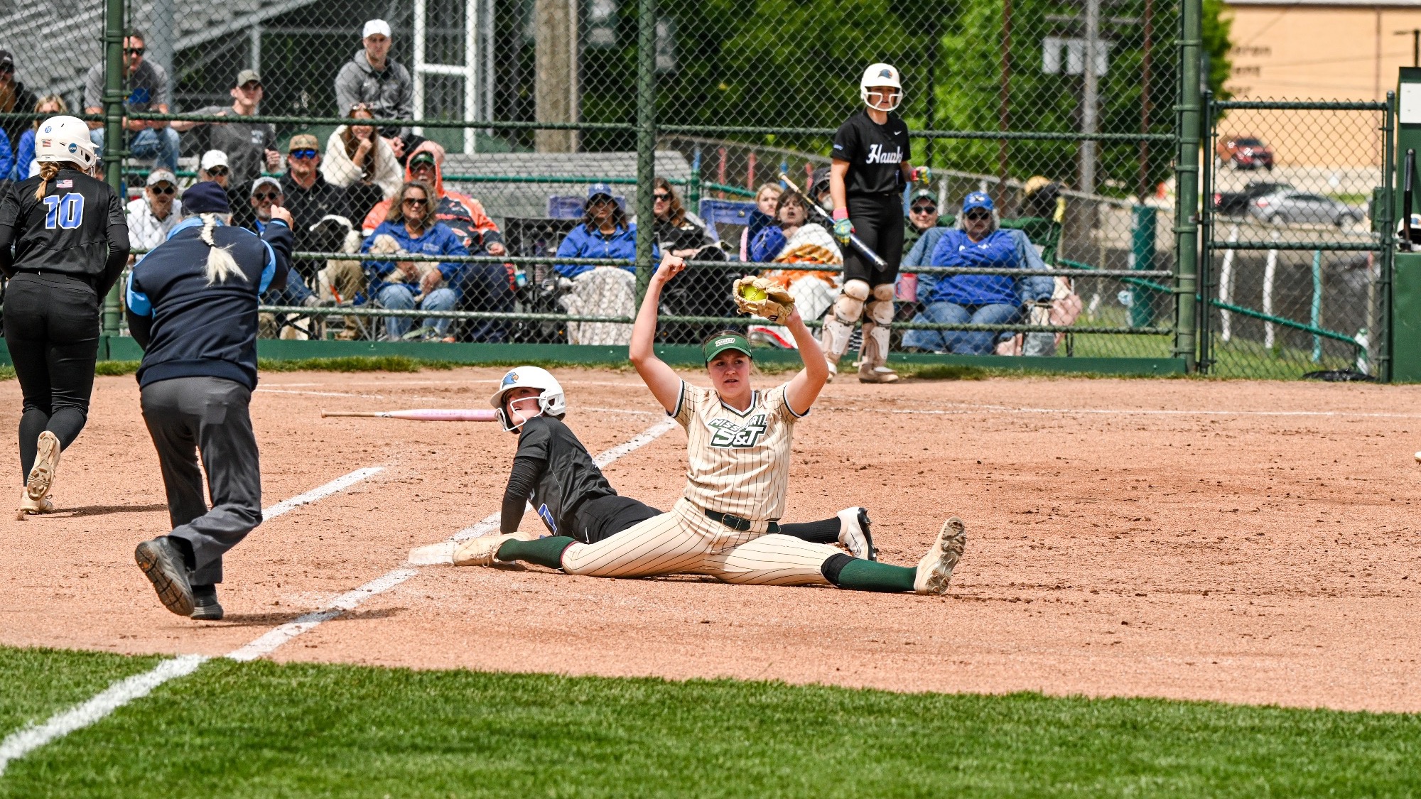 Tori Thompson of Missouri S&T softball, wearing a tan pinstriped uniform with green socks, stretches into a full split at first base to catch a throw, recording an out as a runner slides in, with an umpire and spectators visible at the S&T softball field.