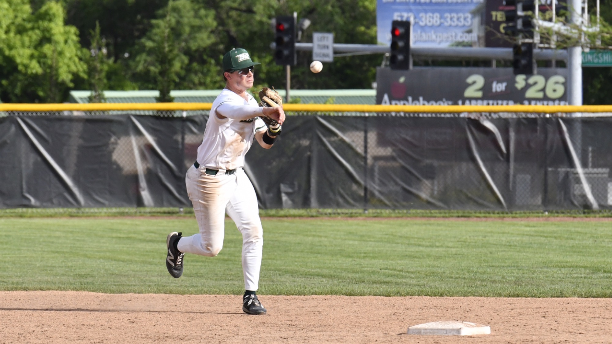 Nick Krodinger of Missouri S&T baseball photographed throwing the baseball from his position at shortstop during a ground out. He wears a white Miners uniform with green trim, a green baseball cap, and sunglasses.