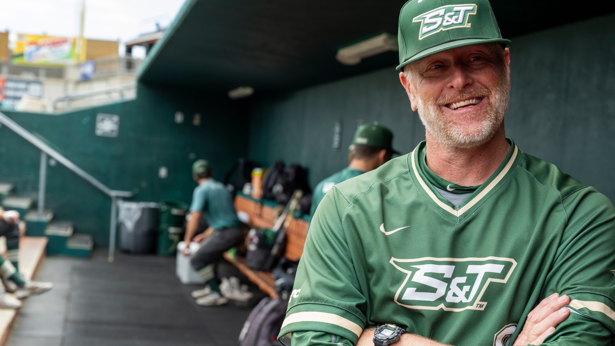 Head baseball coach Todd DeGraffenreid photographed in the Missouri S&T dugout, smiling with arms crossed while wearing a green Miners uniform and cap, as players and equipment are visible in the background.