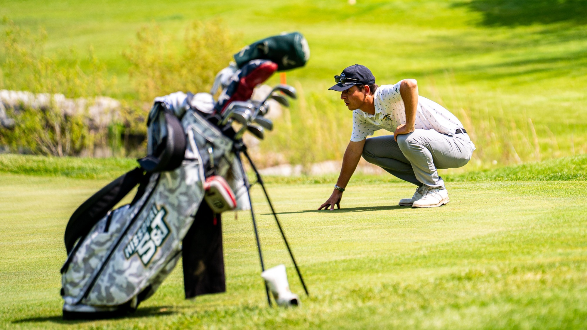 Head Men's Golf Coach Connor Benjamin photographed squatting and examining the green before his athlete putts. He is wearing a white polo with wearing a white polo with gray pants and a black baseball cap. A golf back is visible in the foreground.