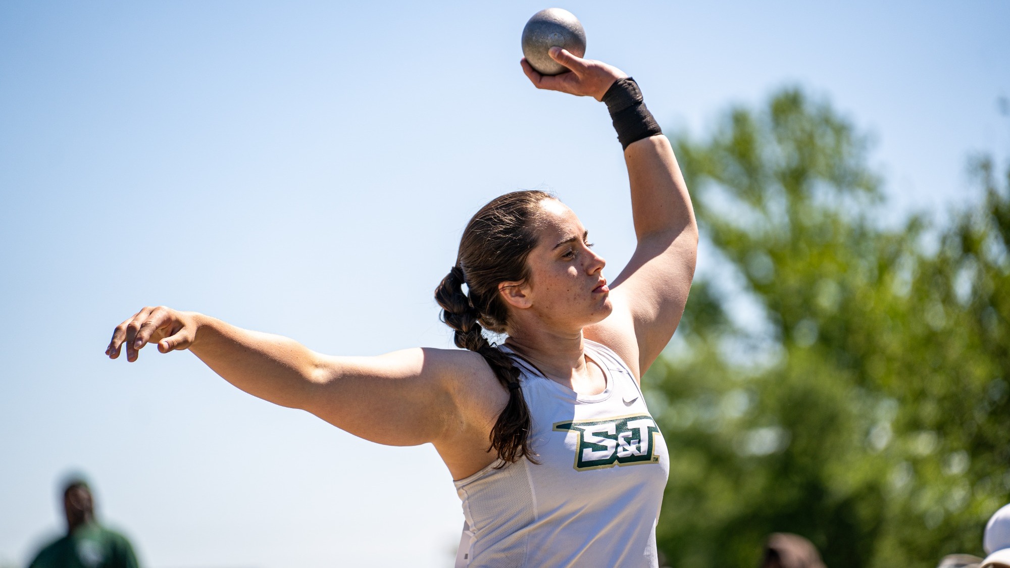 Sydney Morris of Missouri S&T Track and Field photographed holding a shot put above her head as she gets ready to throw. She is wearing a white S&T uniform.
