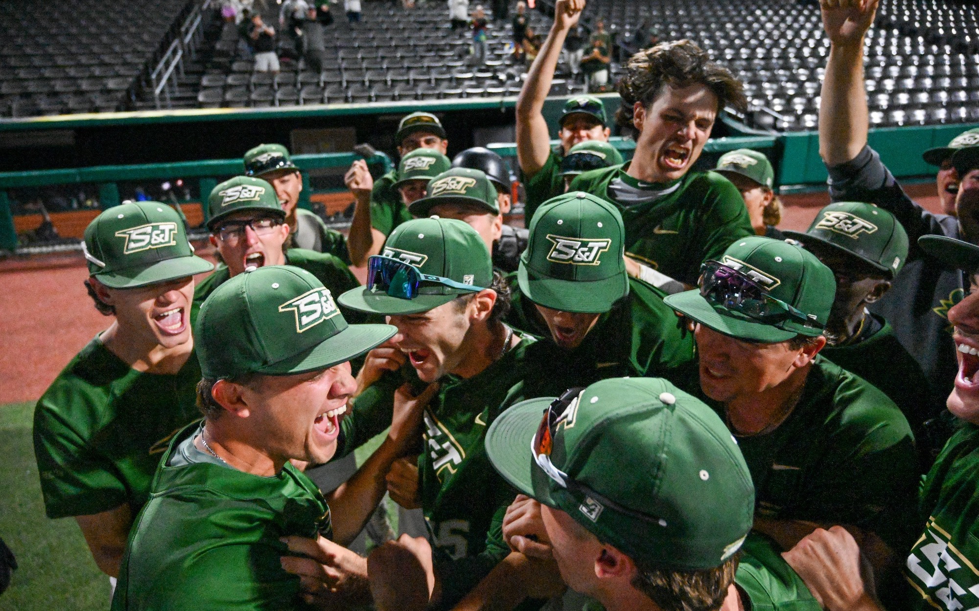 Missouri S&T baseball players celebrate on the field after a comeback win over UIndy in the GLVC semifinals, shouting and raising their arms in excitement as teammates crowd together under the stadium lights.