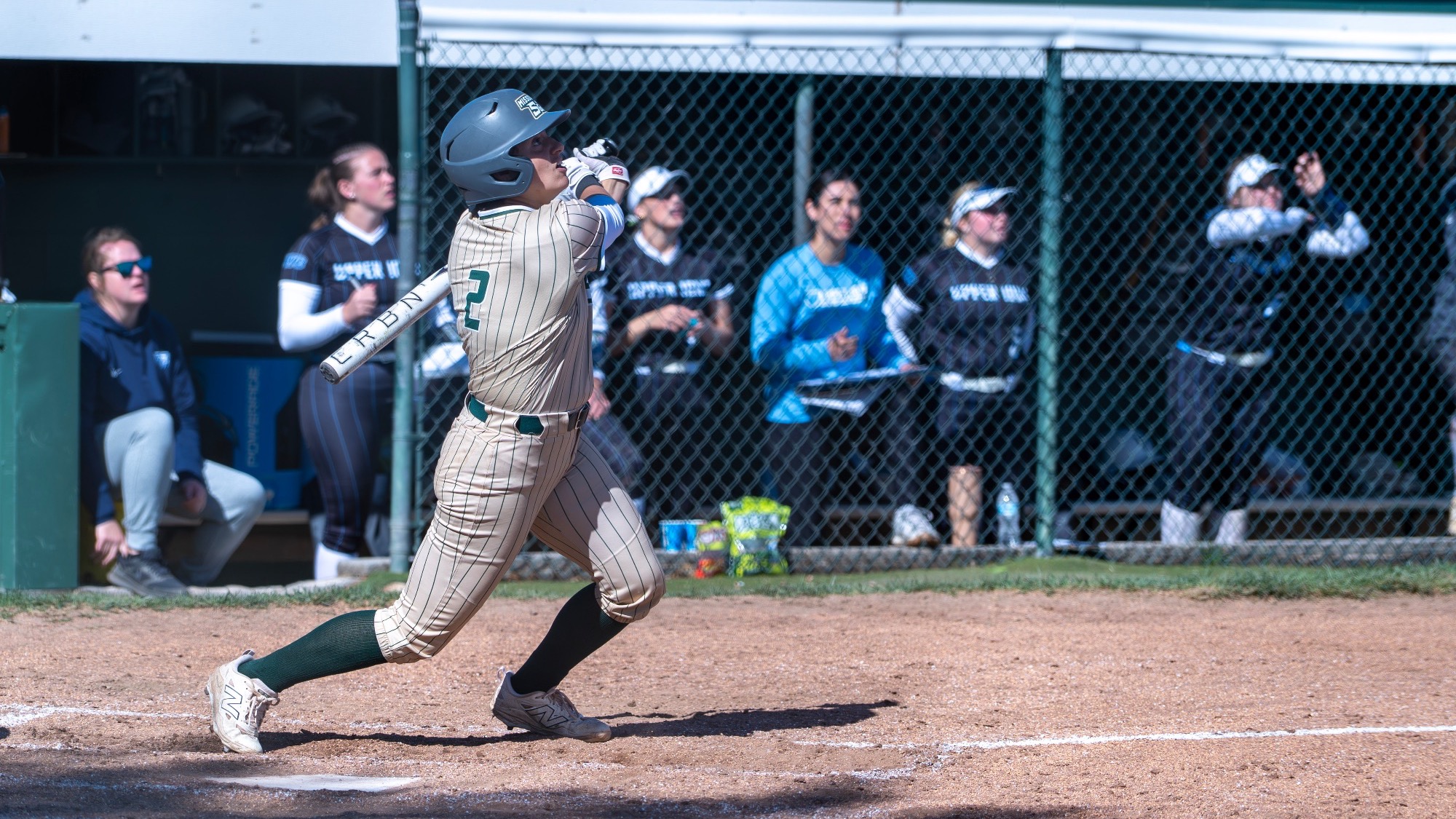 Missouri S&T softball player Taylor Nelson follows through on a swing at the S&T Softball Field. Nelson wears the Miners’ gold pinstripe jersey, matching pants, green socks, and a gray batting helmet while watching the ball off the bat. Opposing players and coaches stand in the Upper Iowa dugout in the background behind the chain-link fence.