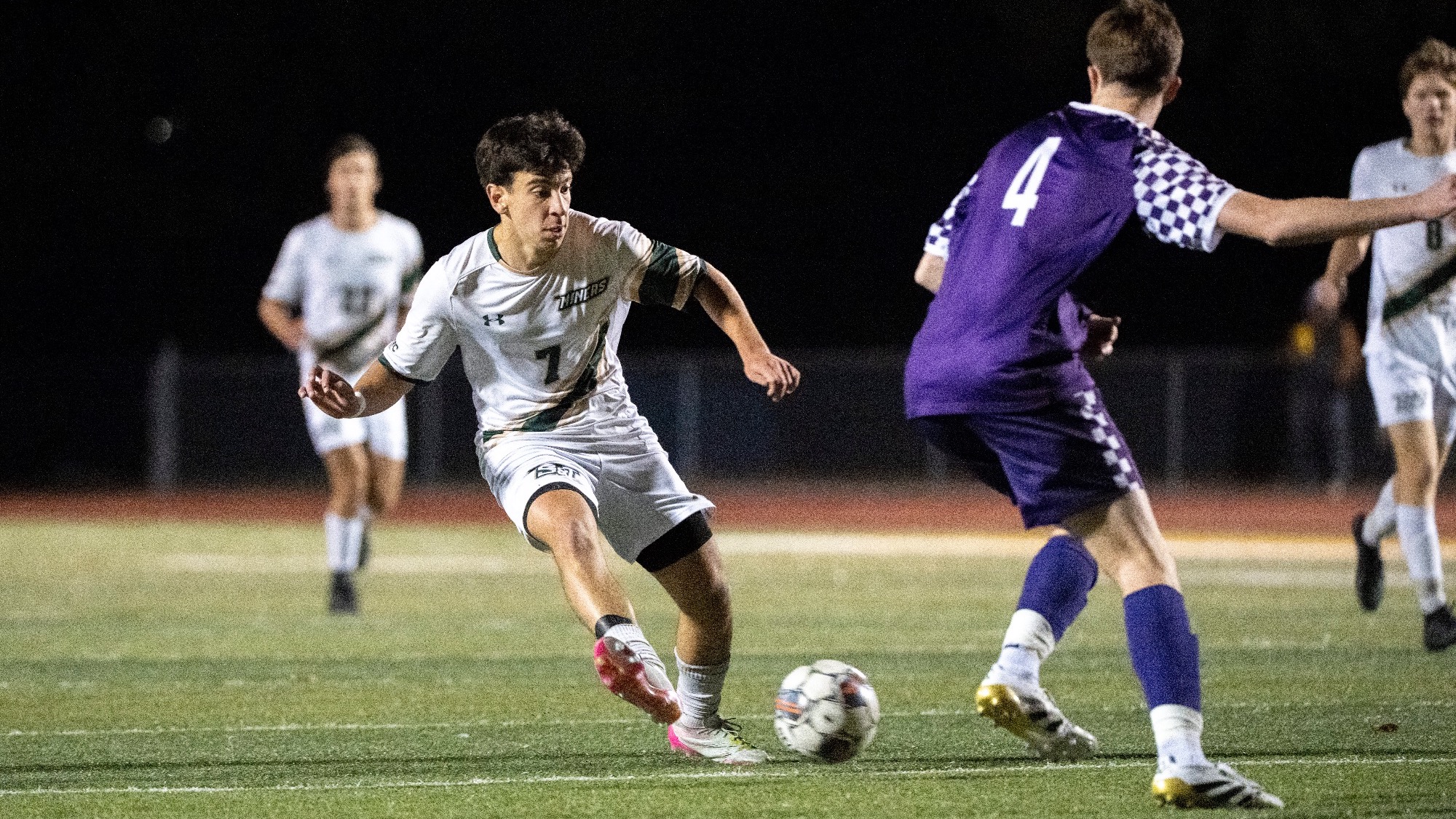 Missouri S&T men's soccer player Joao Vieira (#7) dribbles the ball during a nighttime match against Truman State at Allgood-Bailey Stadium. Vieira wears a white Miners jersey with green accents, white shorts, and white socks as he moves toward a Truman State defender in a purple uniform. Other players are visible in the background under the stadium lights.