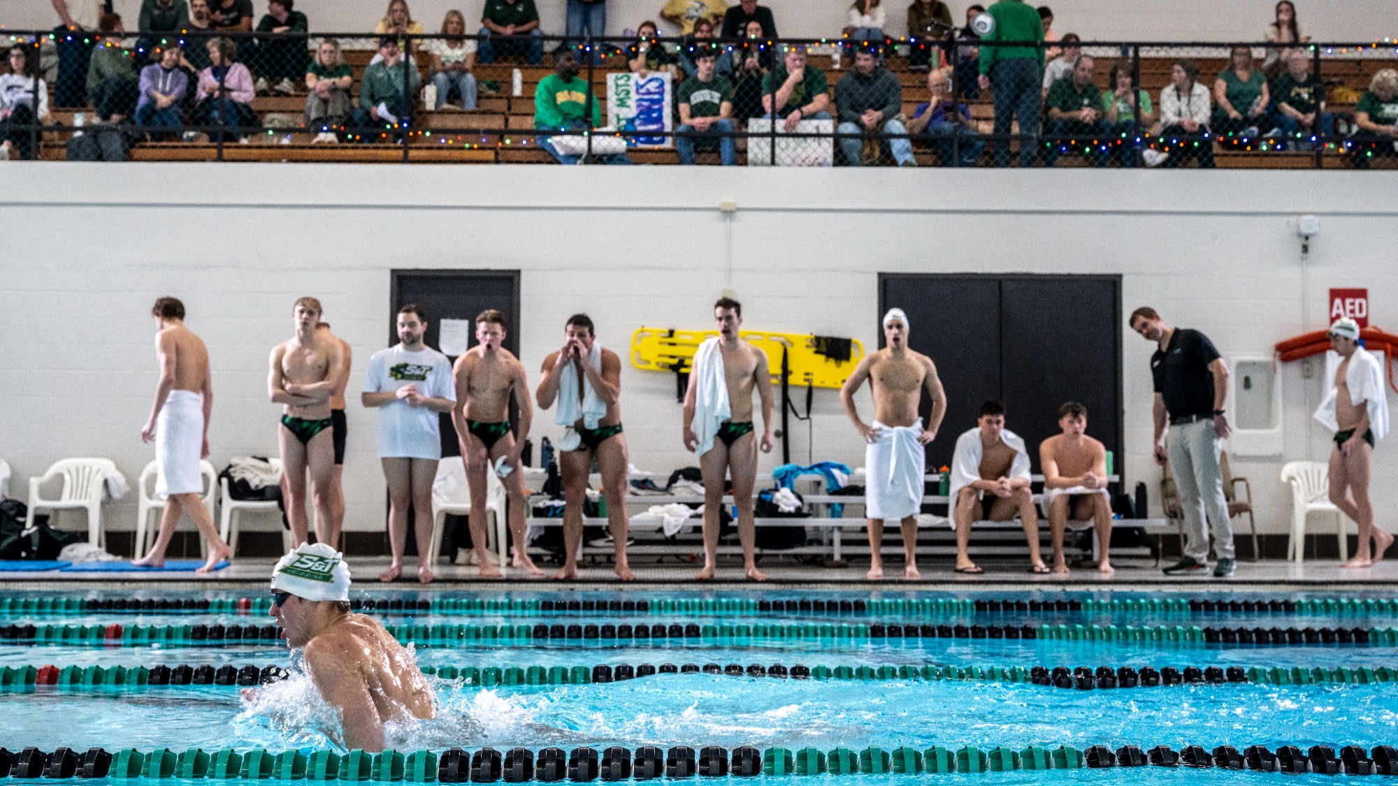 A Missouri S&T swimmer wearing a white team cap competes in a breaststroke race in the foreground of an indoor pool. Along the opposite pool deck, teammates in green-and-black uniforms and coaches watch the race, with several cheering from the deck. Spectators fill the balcony seating above, overlooking the competition.