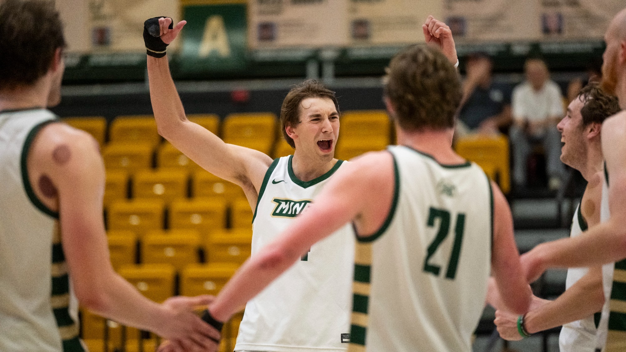 Tyler Johnwick of Missouri S&T Mens volleyball photographed cheering with his fists raised in the air after a point scored by Missouri S&T. He is wearing a white S&T men's volleyball uniform with green trim as teammates gather around him