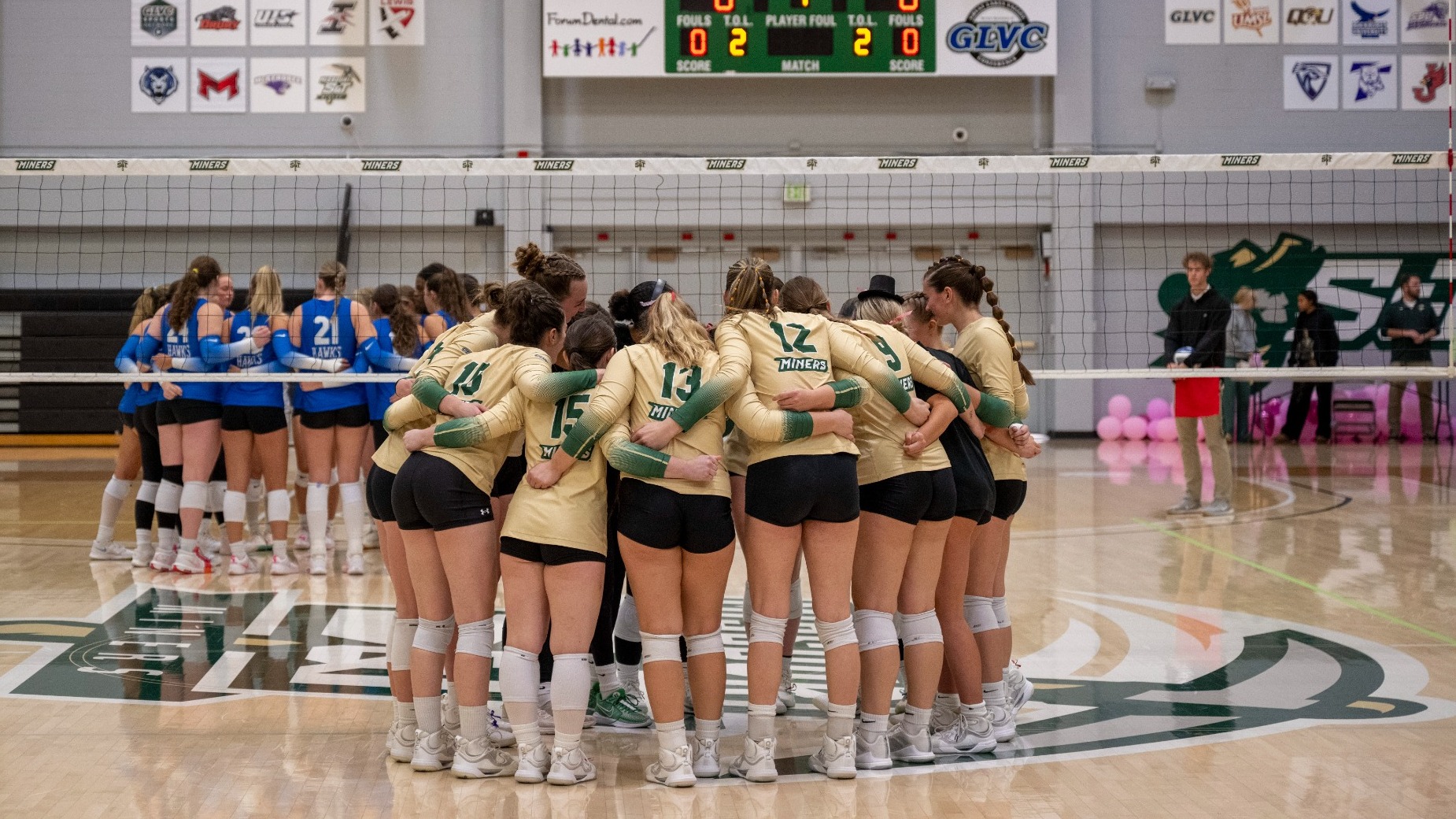 Missouri S&T women’s volleyball players in green and gold uniforms huddle together on the court during a match at Gibson Arena, with the opposing team and scoreboard visible in the background.