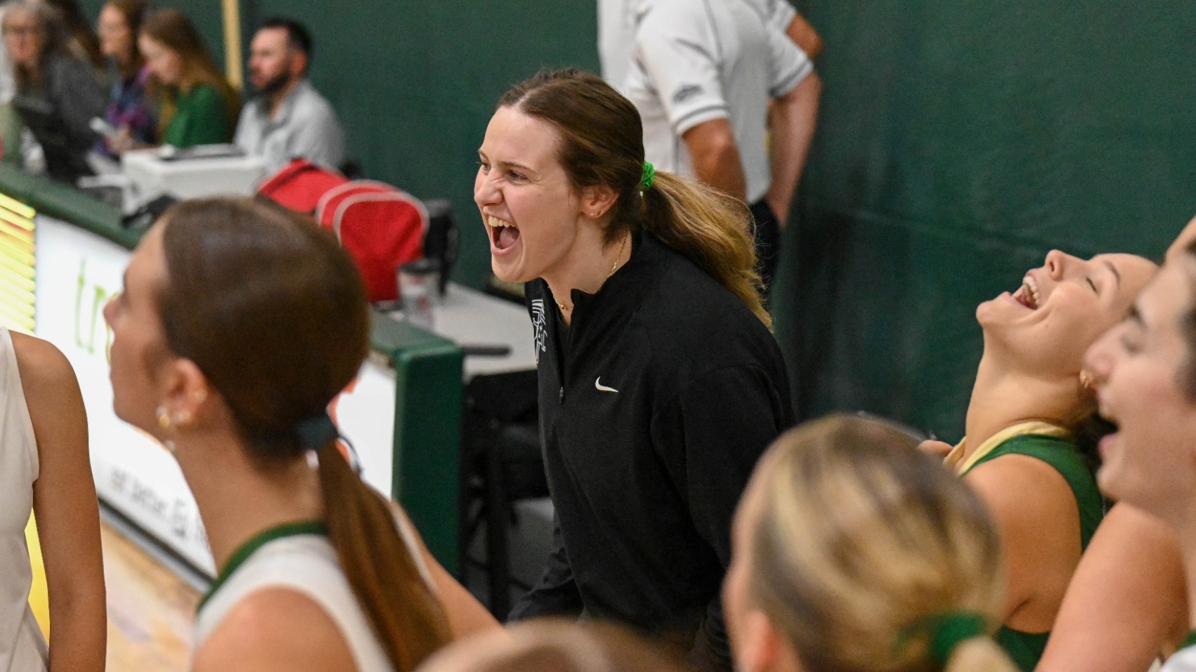 Coach Cooper Martin laughs with Missouri S&T women’s basketball players during a moment on the sideline. Several players in S&T uniforms are gathered around her, smiling and laughing.