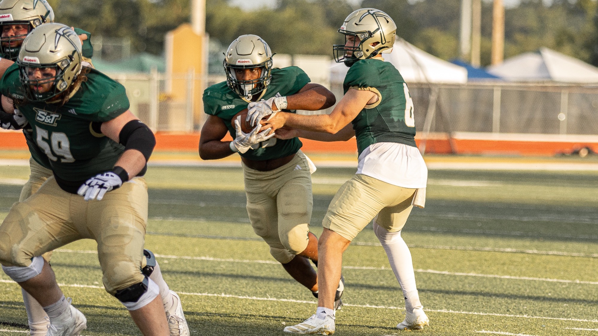 AJ Harris being handed the football by Eli Scott while wearing green uniforms and gold helmets during a football game at Allgood-Bailey stadium