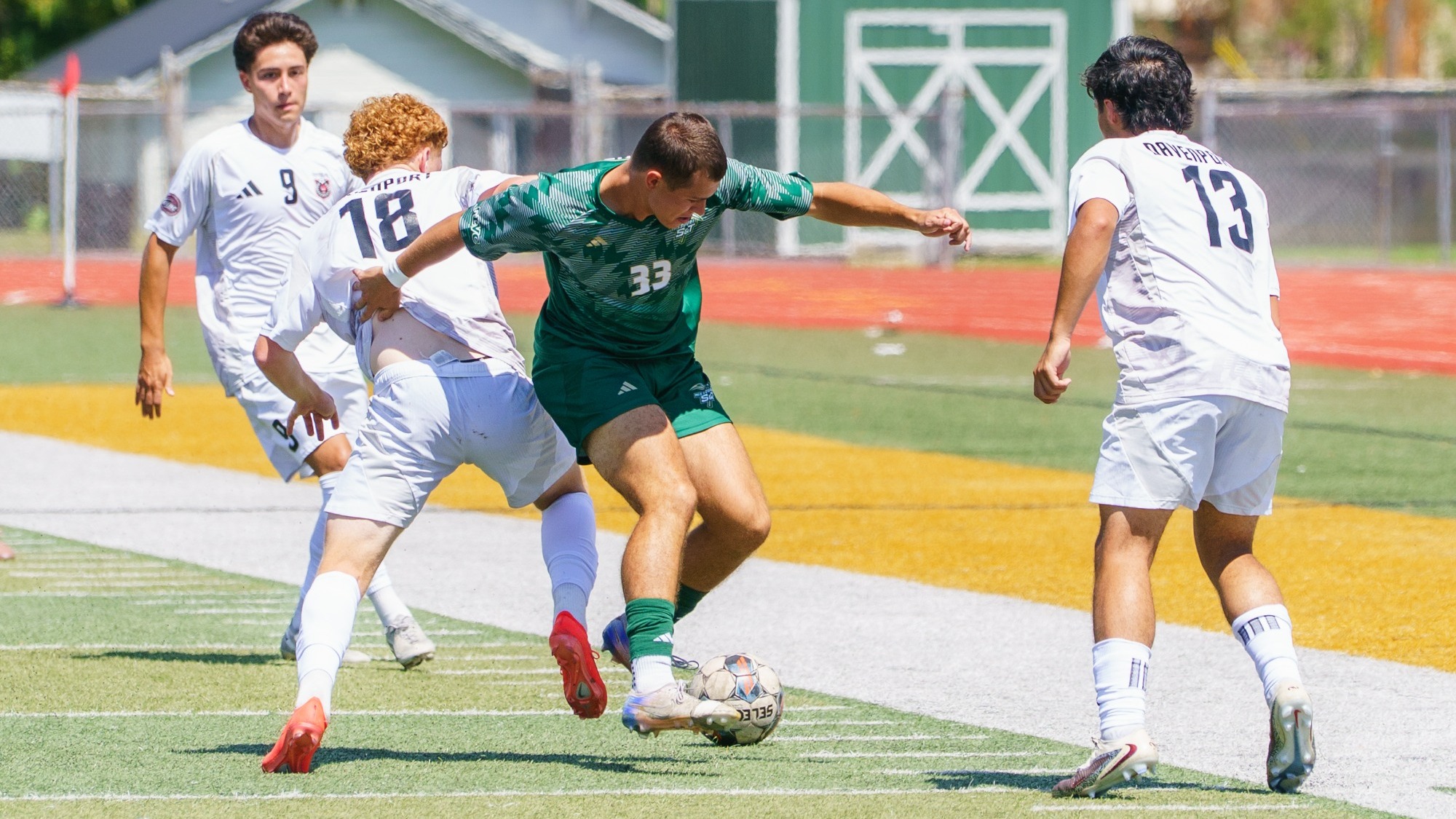 Jack Steele of Missouri S&T Men's Soccer photographed wearing a green kit and dribbling the soccer ball while Davenport defenders wearing white surround him
