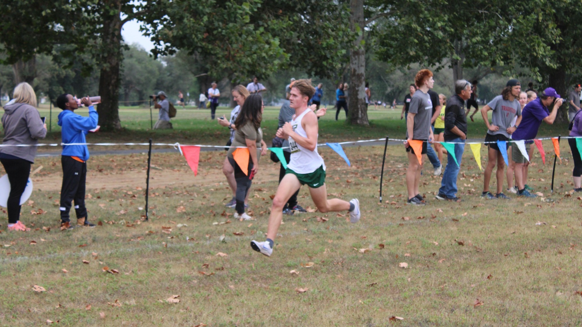 Tate Uding of Missouri S&T Men's Cross Country photographed while running full stride at the Bearcat Invite. He is wearing a white uniform top and green shorts