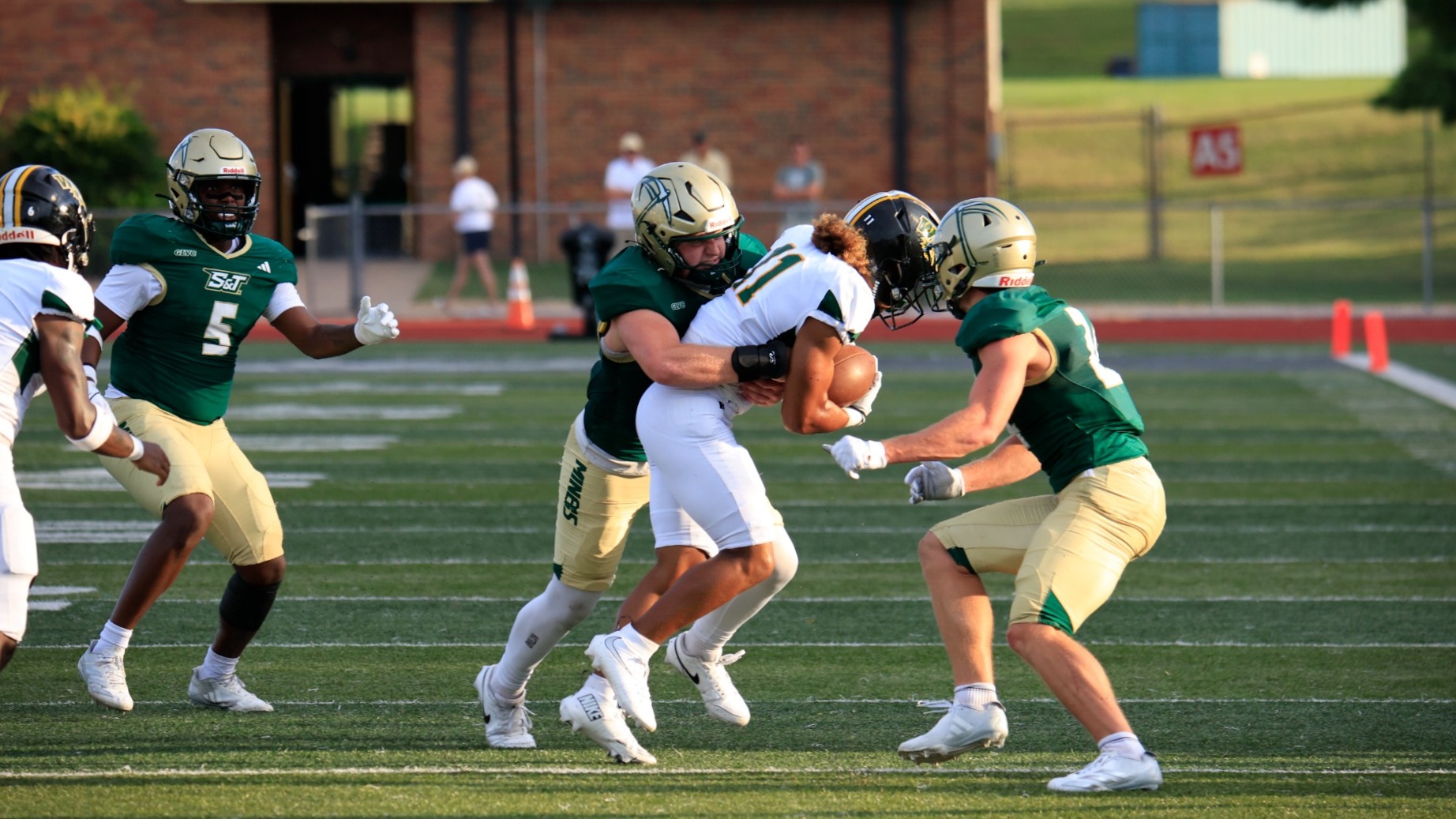 Kaden Glass of Missouri S&T Football photographed tackling a Westgate Christian opponent while a players from both teams are visible around the play. Glass wears a green uniform and gold helmet while the Westgate Christian player wears a white uniform and black helmet.