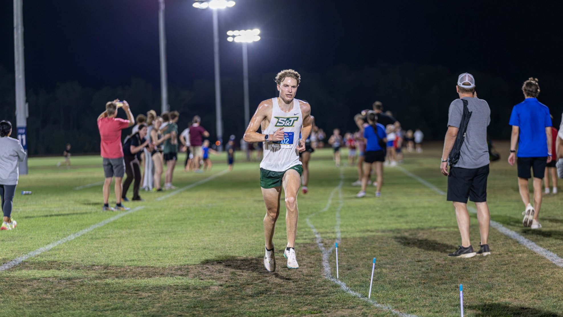 Henry Born of Missouri S&T Cross Country photographed running at night during the Queen City After Dark meet. He is wearing a white running top with green runners shorts and a racing bib