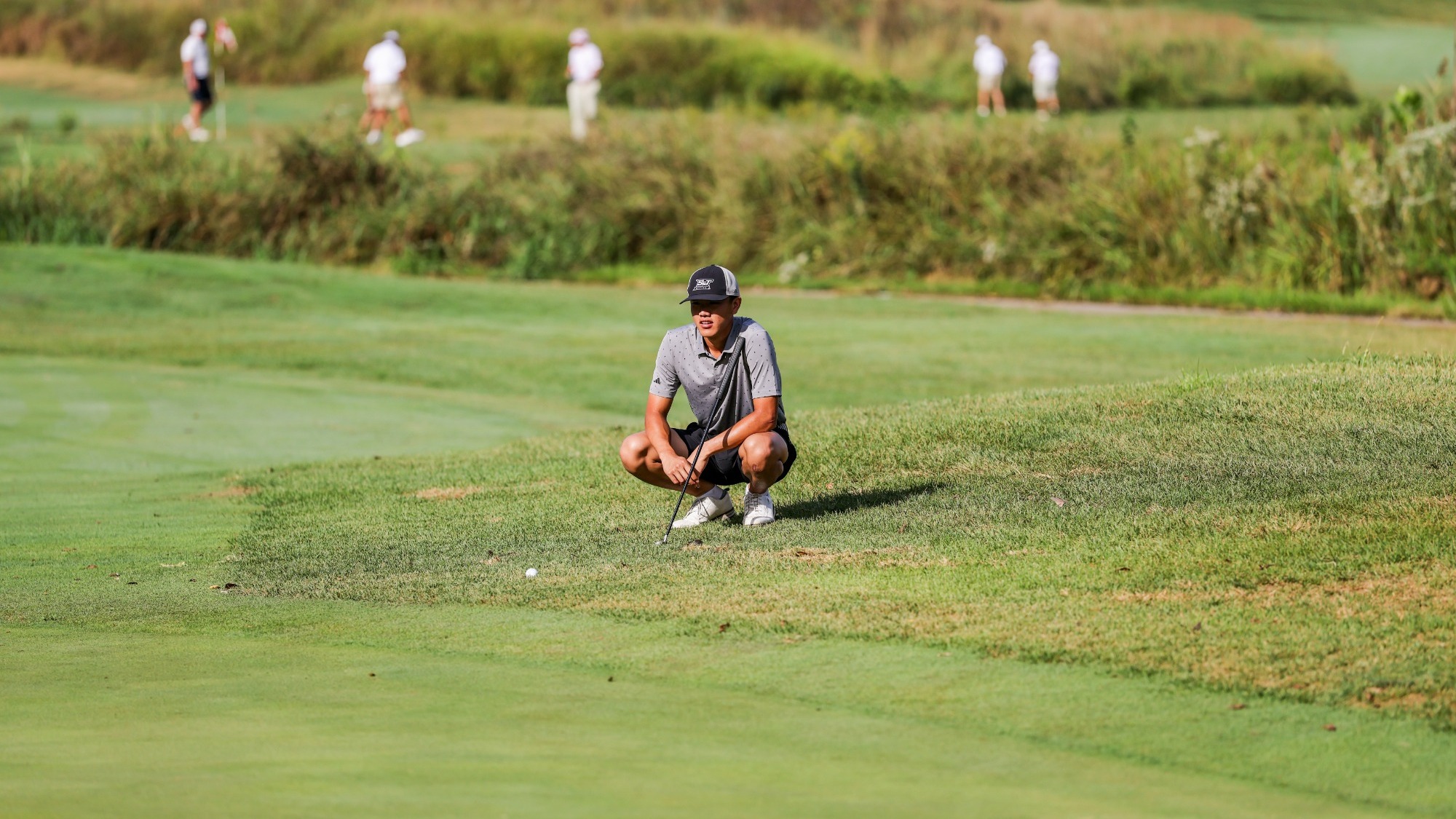 Andy Su of Missouri S&T Golf photographed wearing a gray top and black shorts and baseball cap. He is squatting on the golf course to evaluate his next shot.