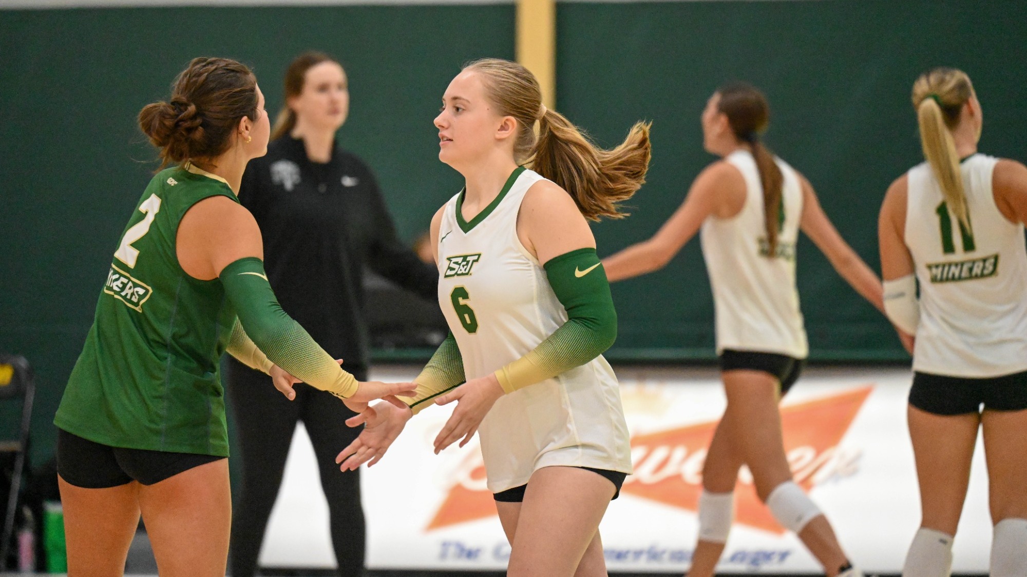 Kaitlyn Brady of Missouri S&T Women's Volleyball photographed wearing a white uniform and low fiving a teammate as she goes back to serve during a match at MSSU
