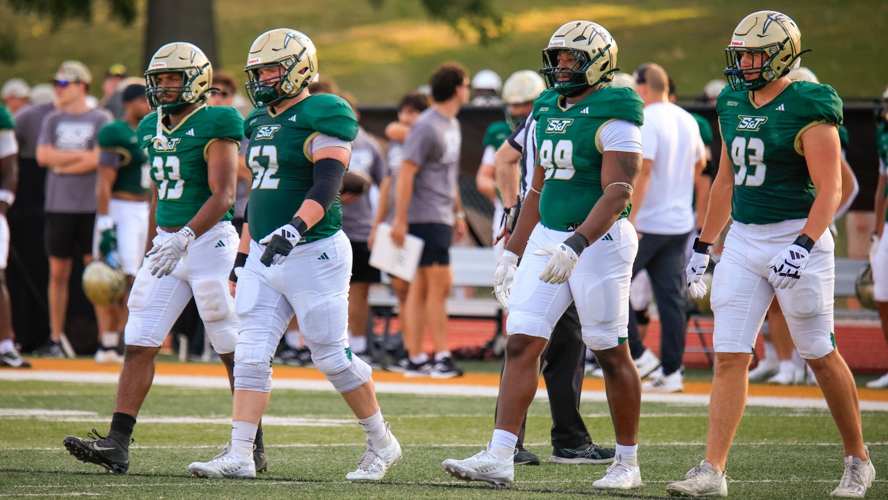 Four Missouri S&T football players — AJ Harris, Thomas Schulte, Christian Kemp and Wes Mayo — walk across the field in green S&T jerseys, white football pants and helmets during a game. Harris, Schulte, Kemp and Mayo are shown from left to right, with teammates, coaches and spectators visible in the background.