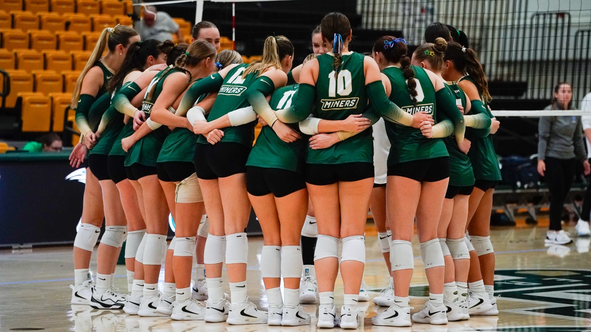Women's Volleyball Team photographed huddling before a match in Gibson. They are all wearing Green uniforms