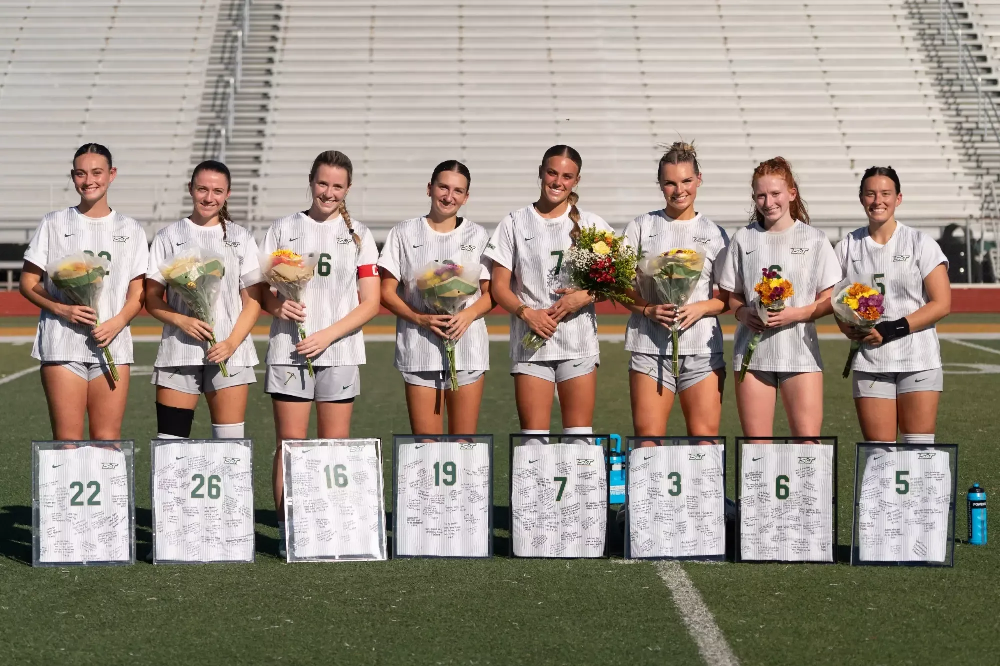 All eight women's soccer Seniors standing in white jerseys and white shorts holding their cased jerseys and flowers all standing next to each other
