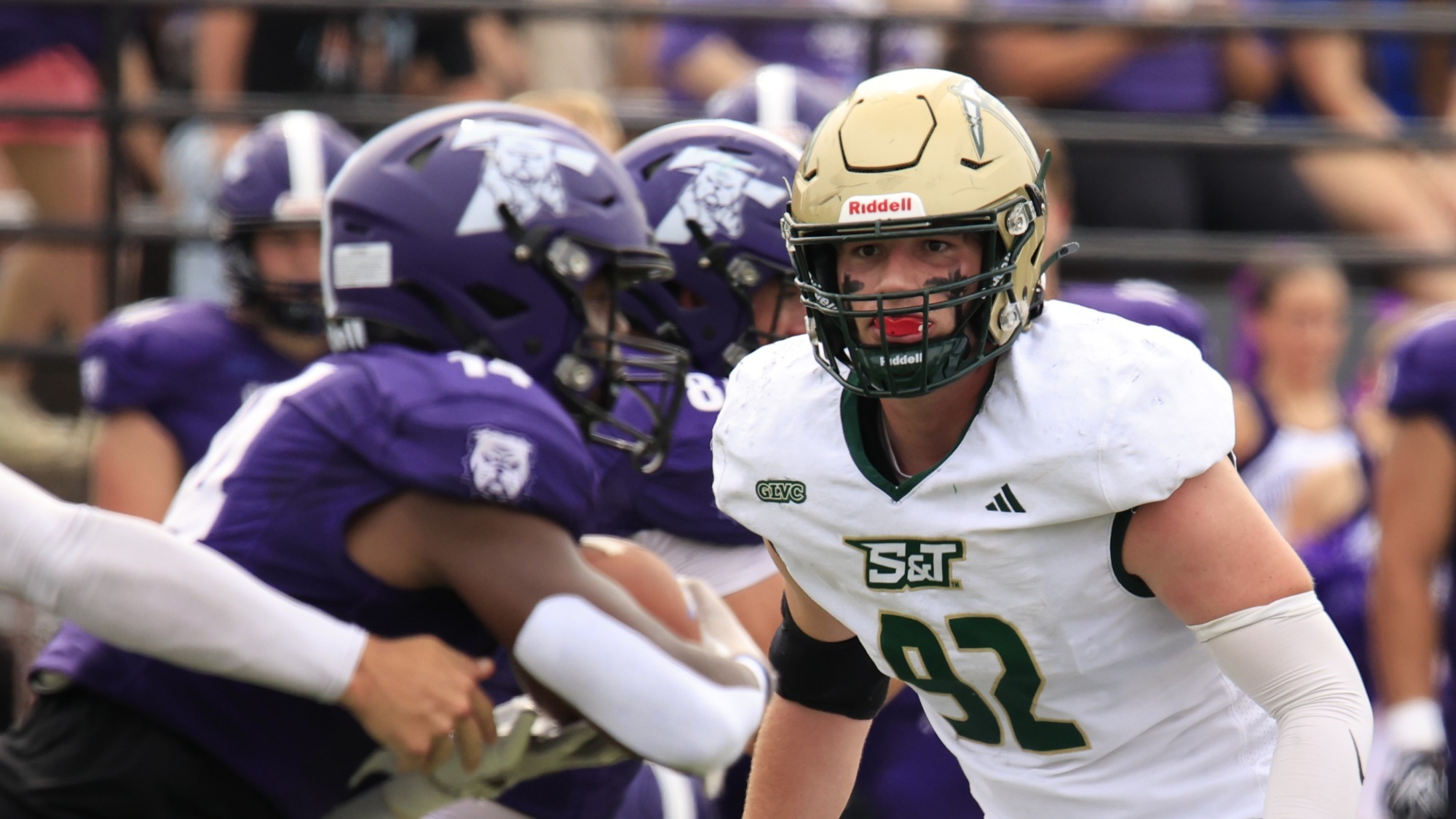 Louis Traglia of Missouri S&T football photographed wearing a white jersey and gold helmet. He watches an opponent wearing a purple jersey and purple helmet receive a handoff during a game in Kirksville, Missouri