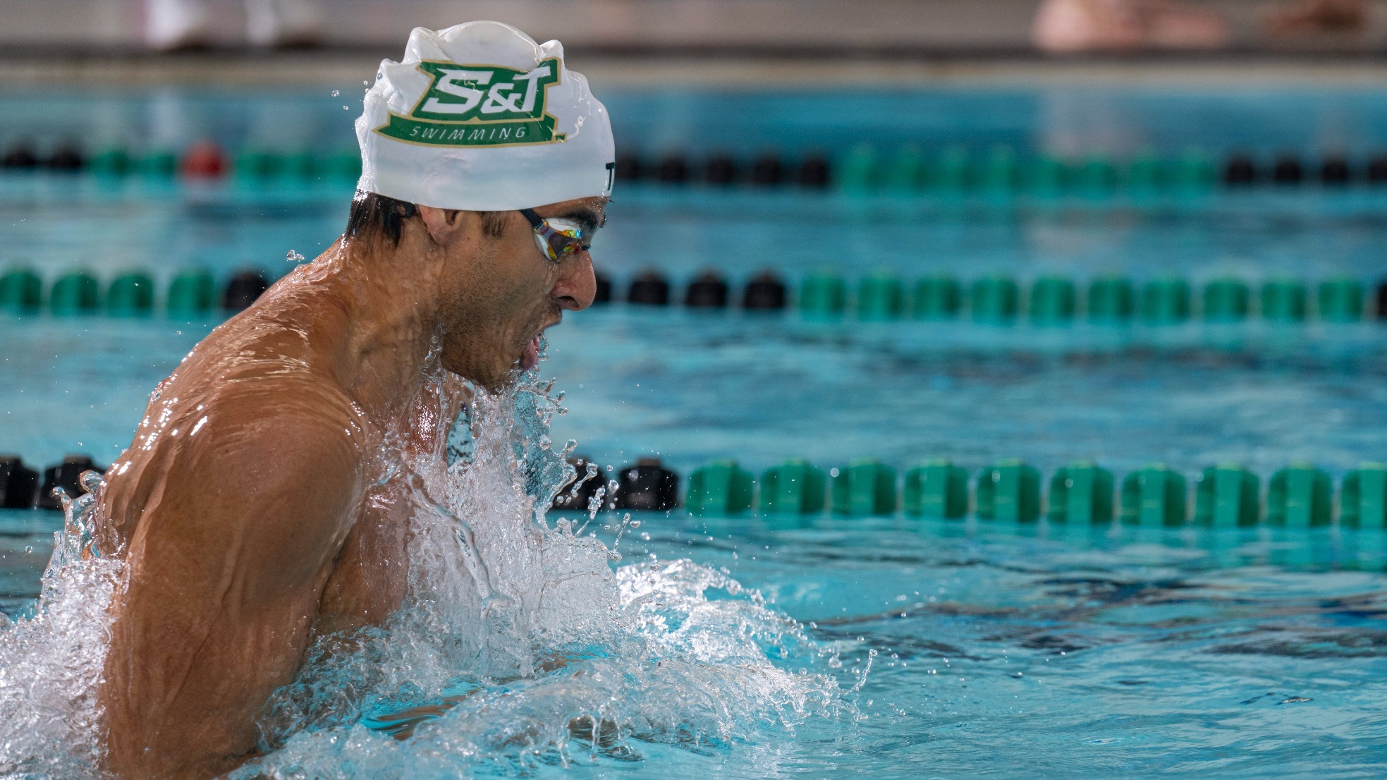 Artur Becker, a Missouri S&T swimmer, competes in a race, wearing an S&T swim cap and goggles as water splashes around him in the pool.